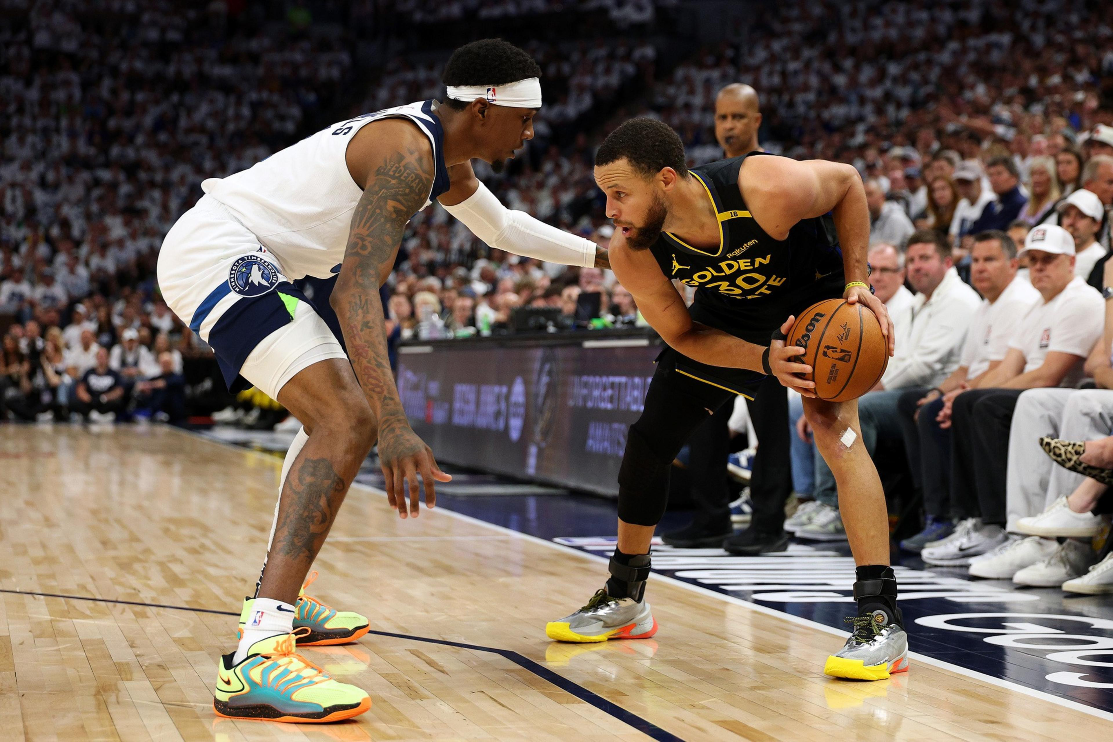 Two basketball players, one in a black jersey holding the ball, the other in white defending closely. A crowd watches intently from the stands.