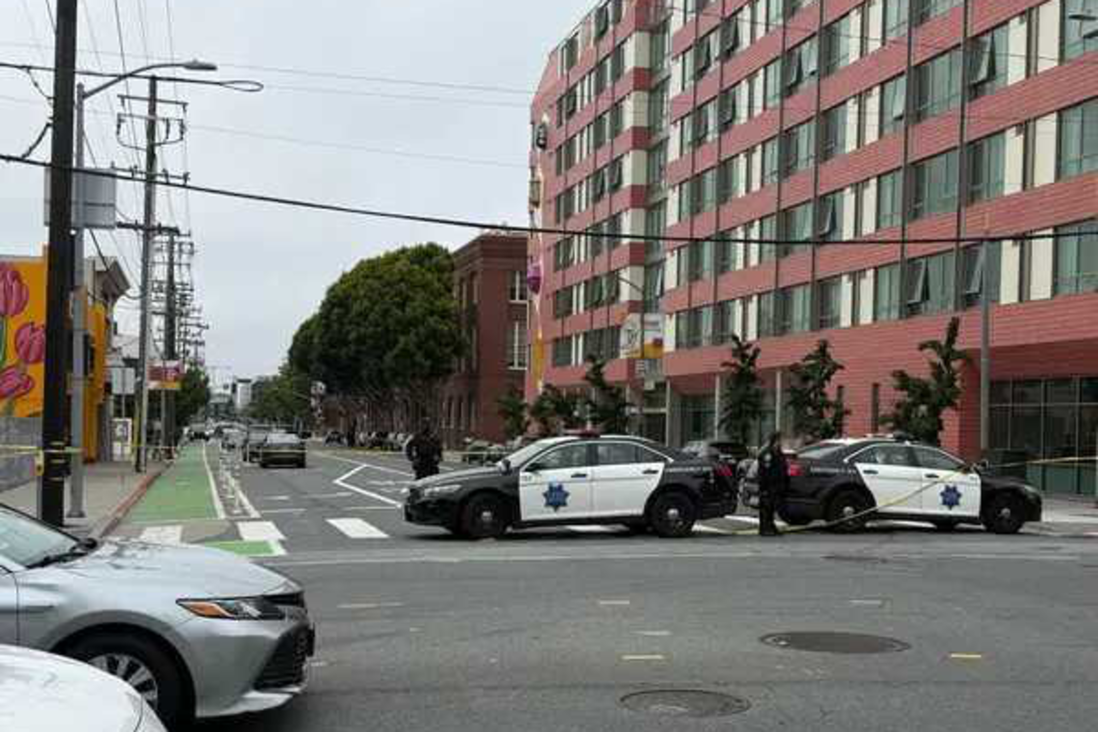 Two police cars block a street near a large red and white apartment building, with officers standing outside and a green bike lane in the foreground.