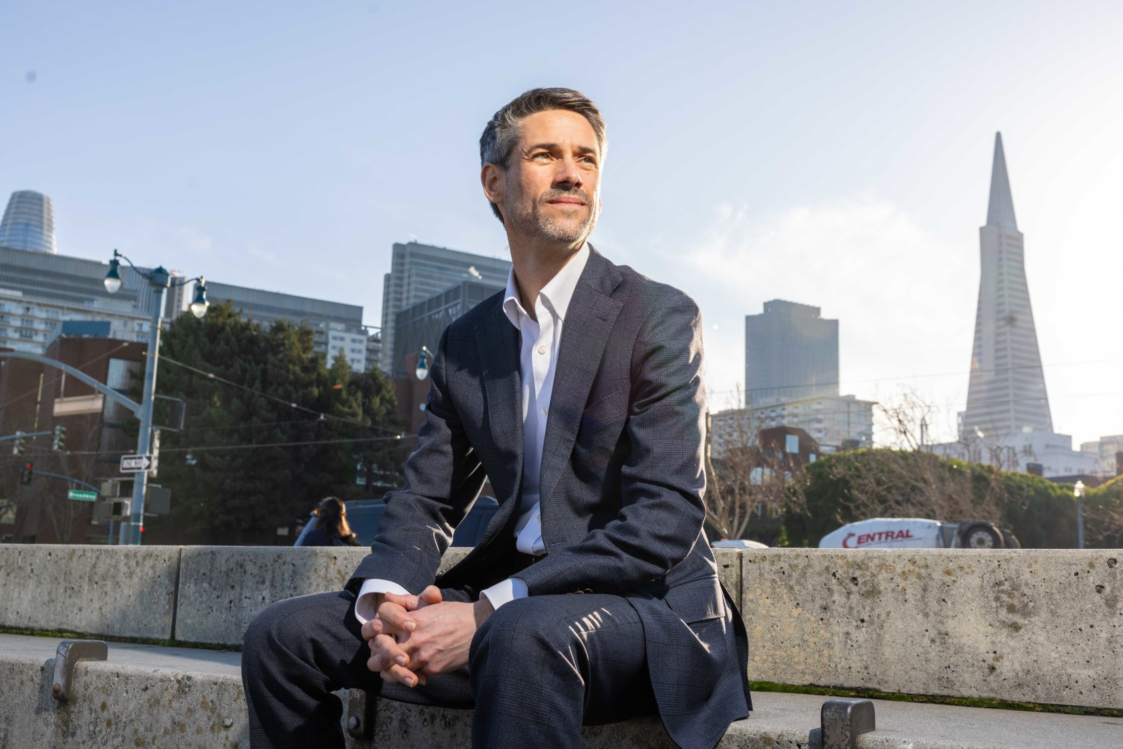 A man in a dark suit sits on concrete steps, looking thoughtfully to the side with a city skyline and the Transamerica Pyramid in the background.