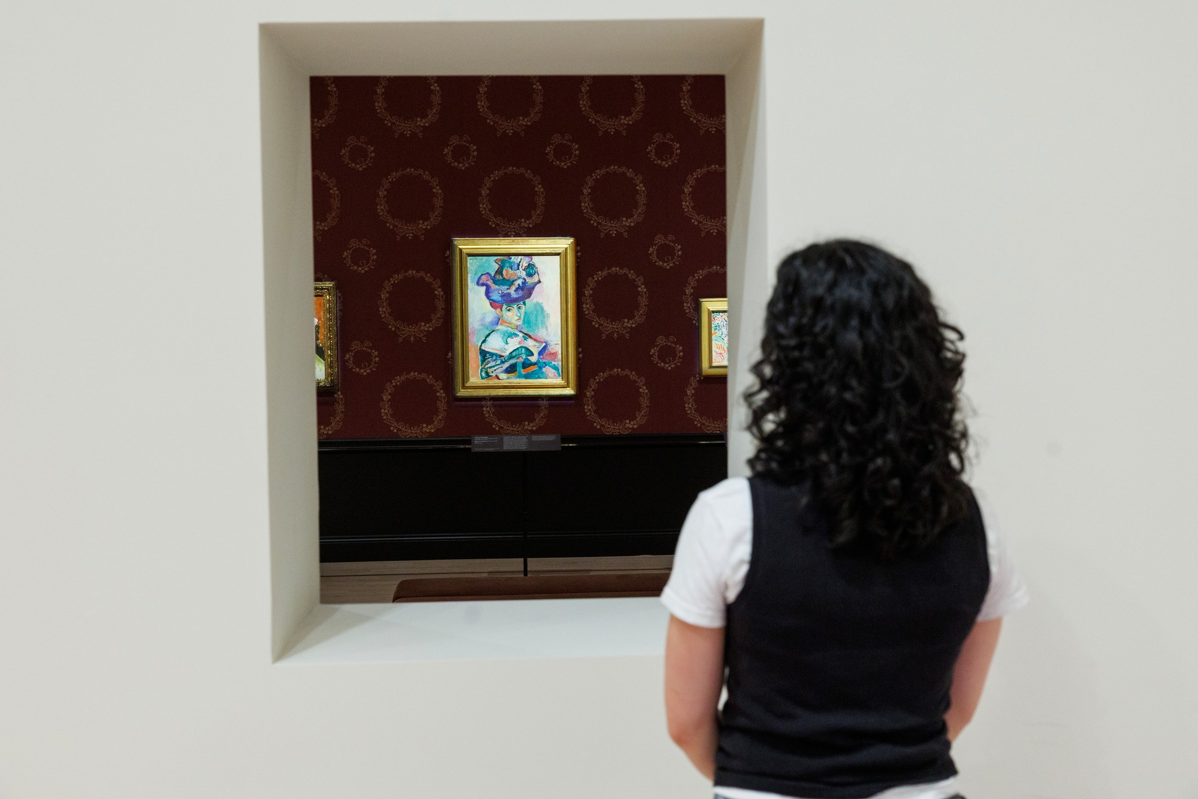 A person with curly dark hair wearing a black vest and white shirt views colorful framed paintings through a square window in a beige wall.