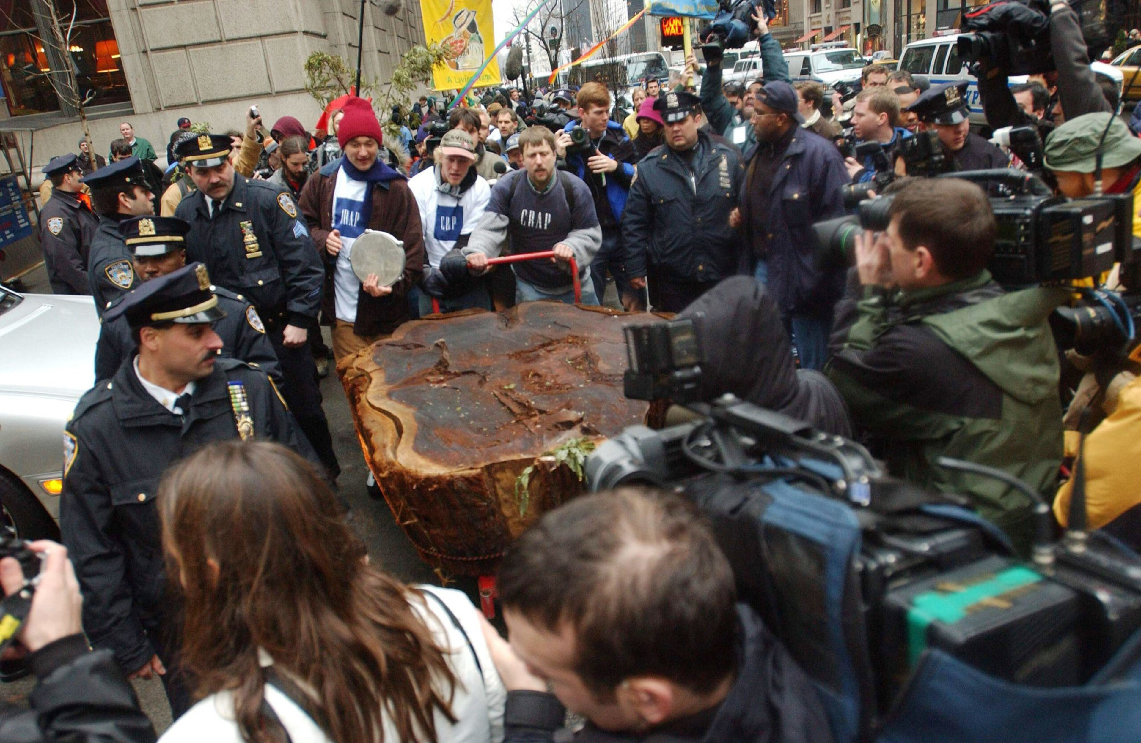 A group of police officers and people crowd around a large tree stump on a red cart amid photographers capturing the scene.