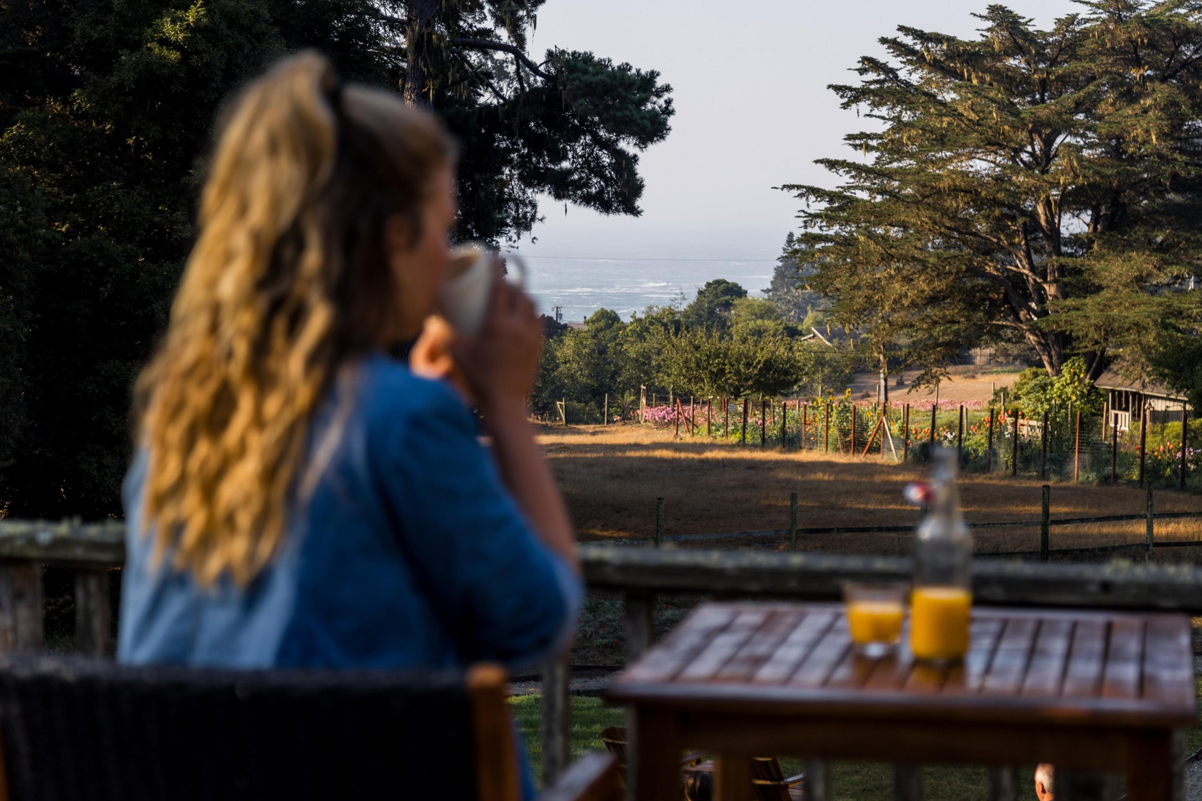 A person with long, wavy hair sits outdoors, sipping a beverage. A table with two glasses of orange juice and a water bottle is in the foreground, and a scenic landscape stretches out behind them.