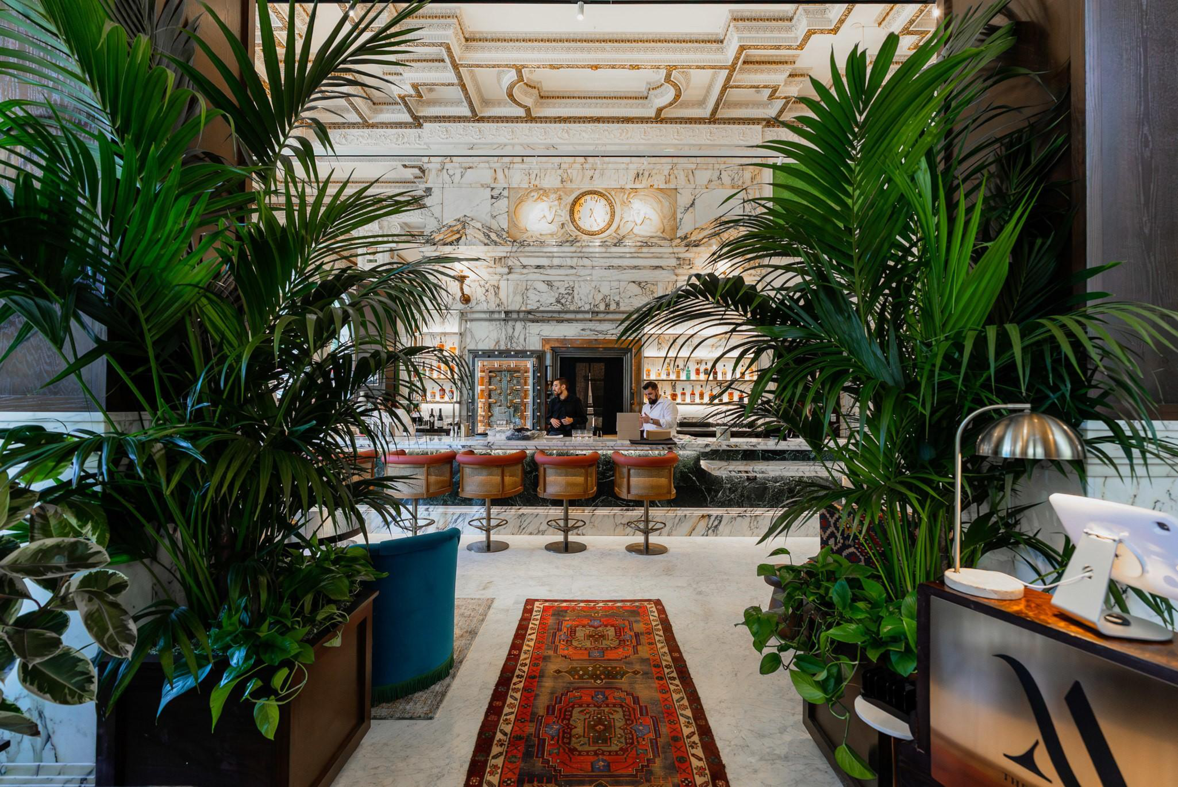 A marble bar with five stools has two bartenders behind it, surrounded by lush green plants and a decorative red patterned rug on the floor.