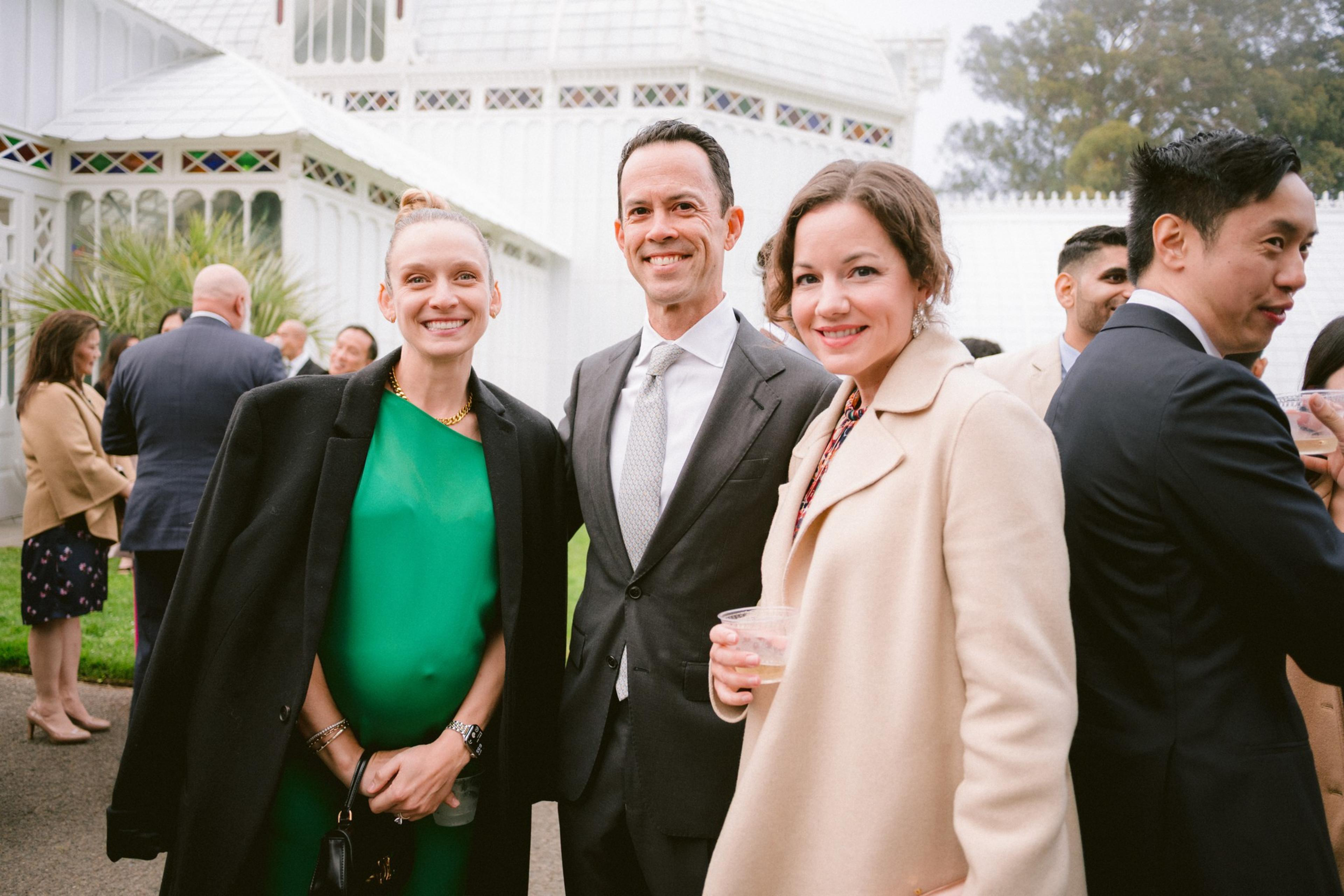 Three people dressed in formal attire smile at the camera outdoors, with a white building and more guests visible in the background.