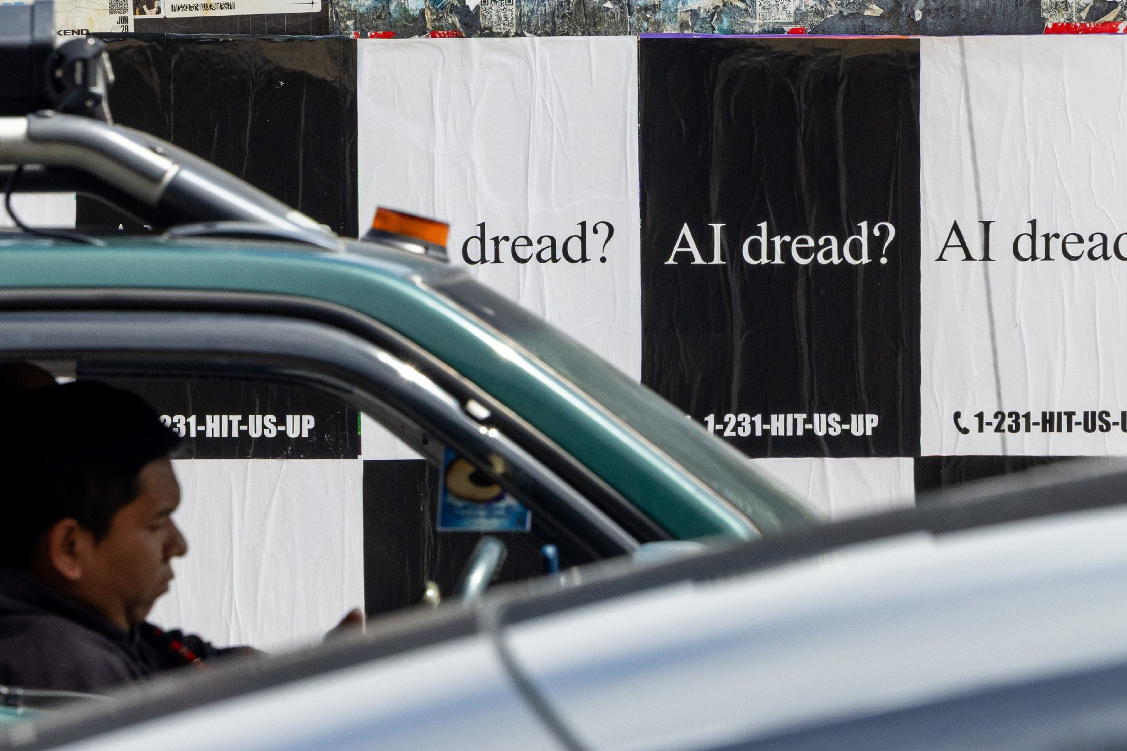 A man sits inside a green car with a roof light, while a wall behind displays black and white posters reading “AI dread?” and the phone number “1-231-HIT-US-UP.”