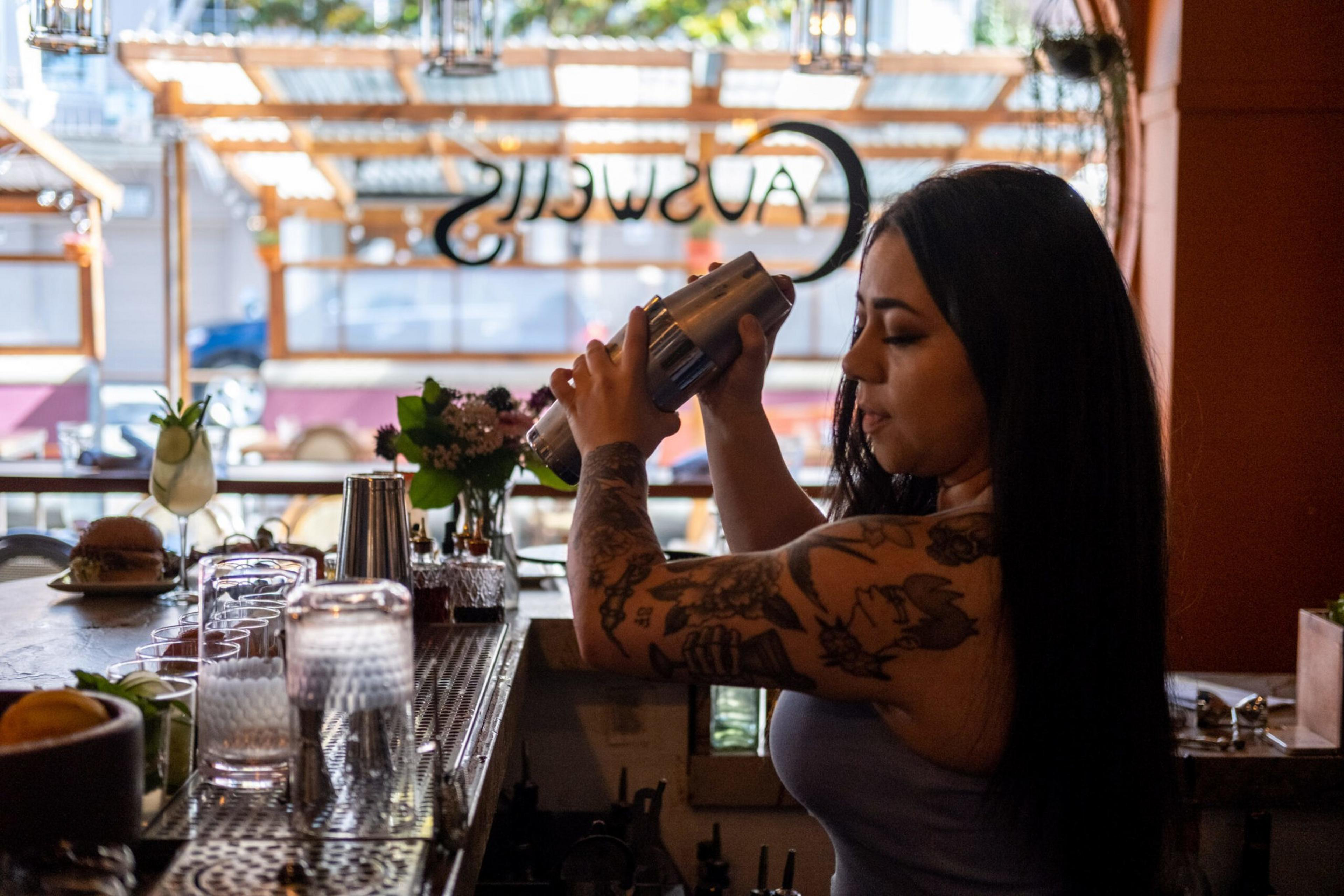 A tattooed woman with long black hair shakes a cocktail shaker behind a bar with glasses and bottles on the counter in a cozy, sunlit setting.