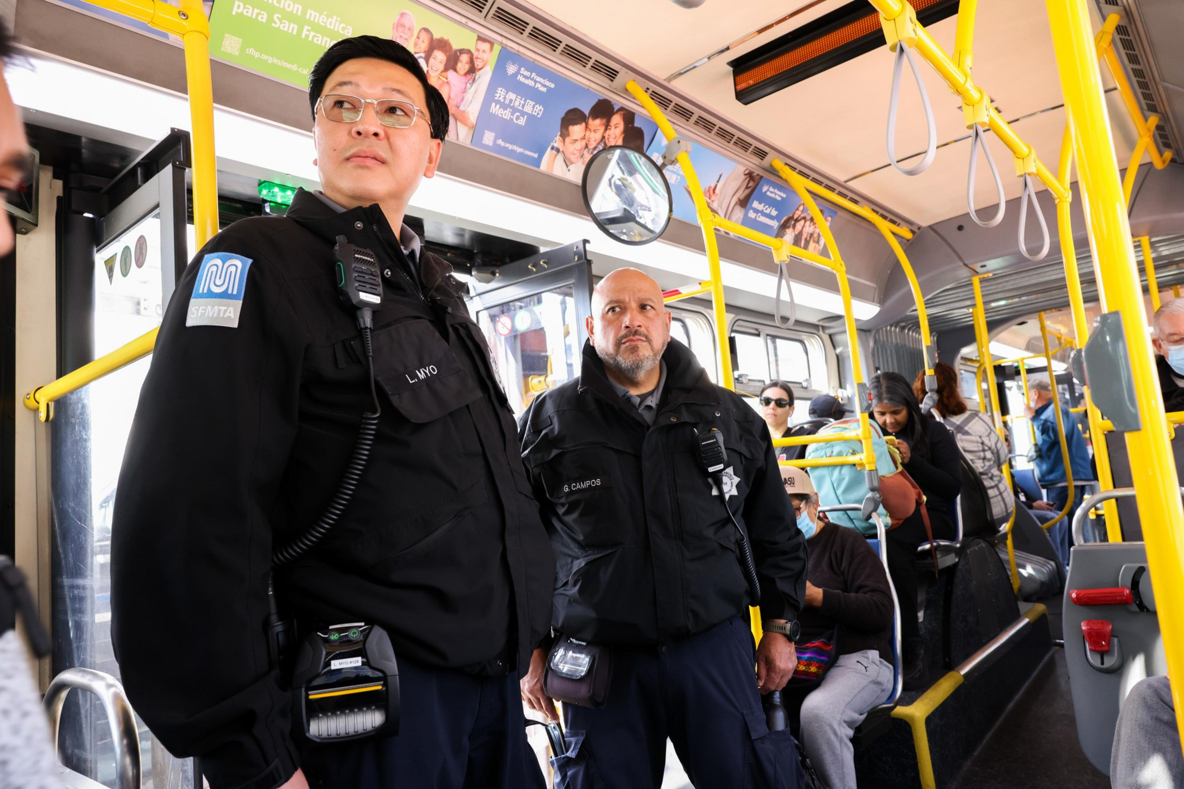 Two uniformed officers are standing on a bus, surrounded by seated and standing passengers. Bright yellow handrails and overhead ads are visible.