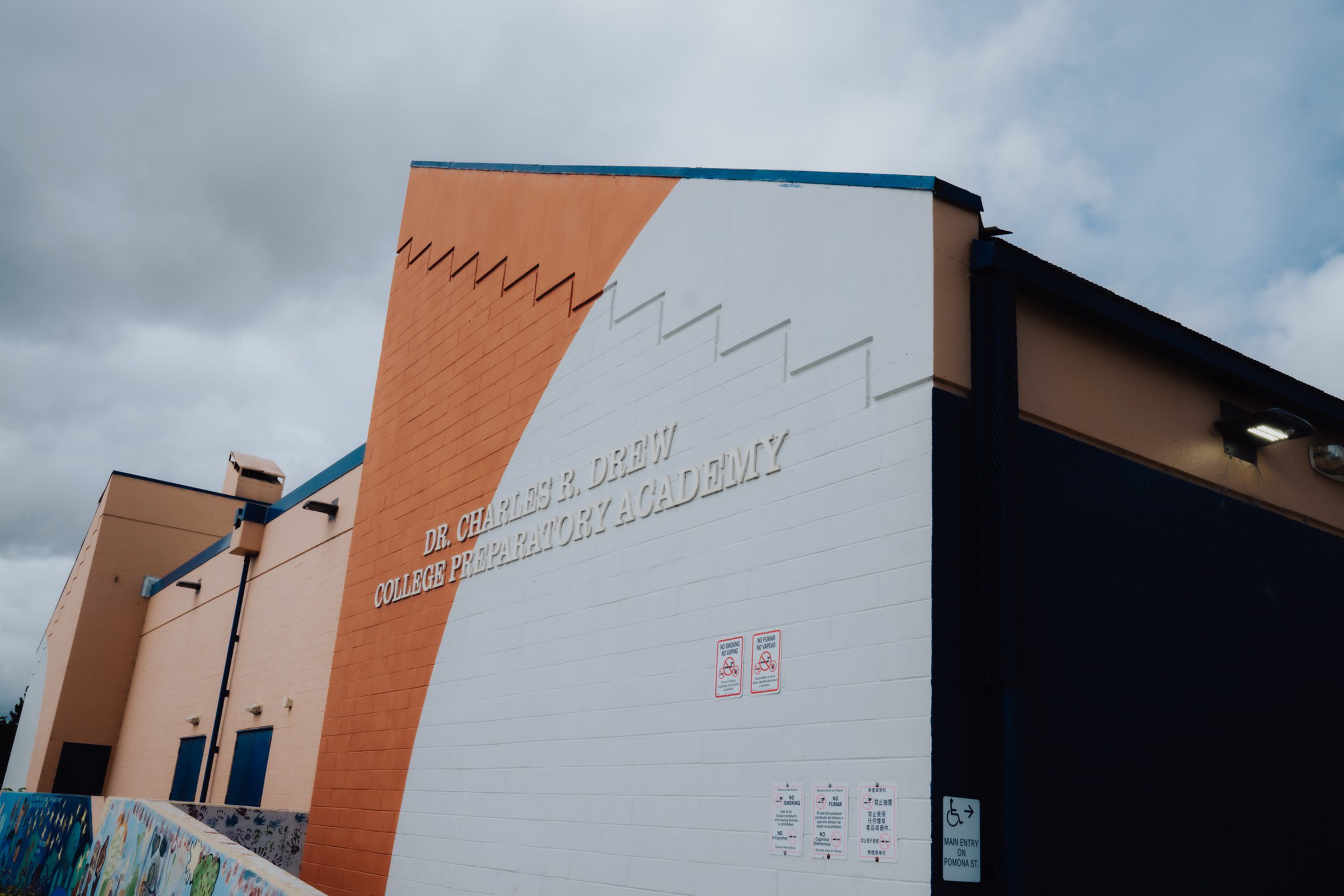A building featuring a large orange and white wall with “Dr. Charles R. Drew College Preparatory Academy” written on it, under a cloudy sky.