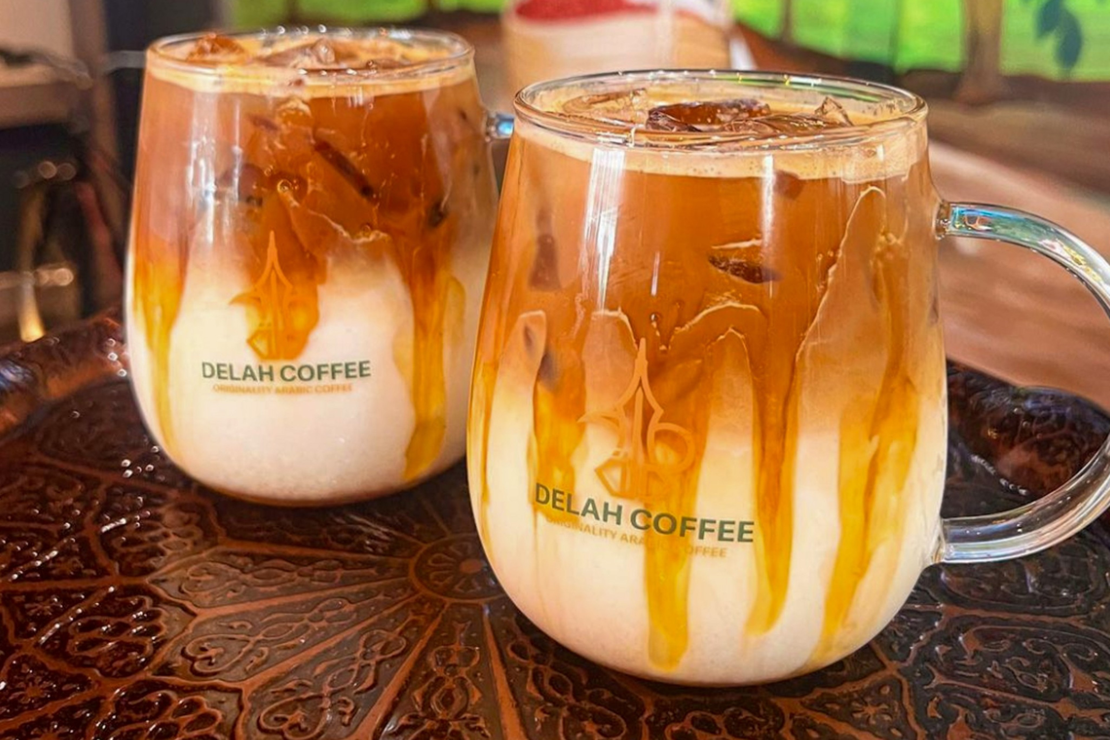 Two glass mugs of iced coffee with milk swirling through, placed on a decorative brown tray, each labeled "DELAH COFFEE."