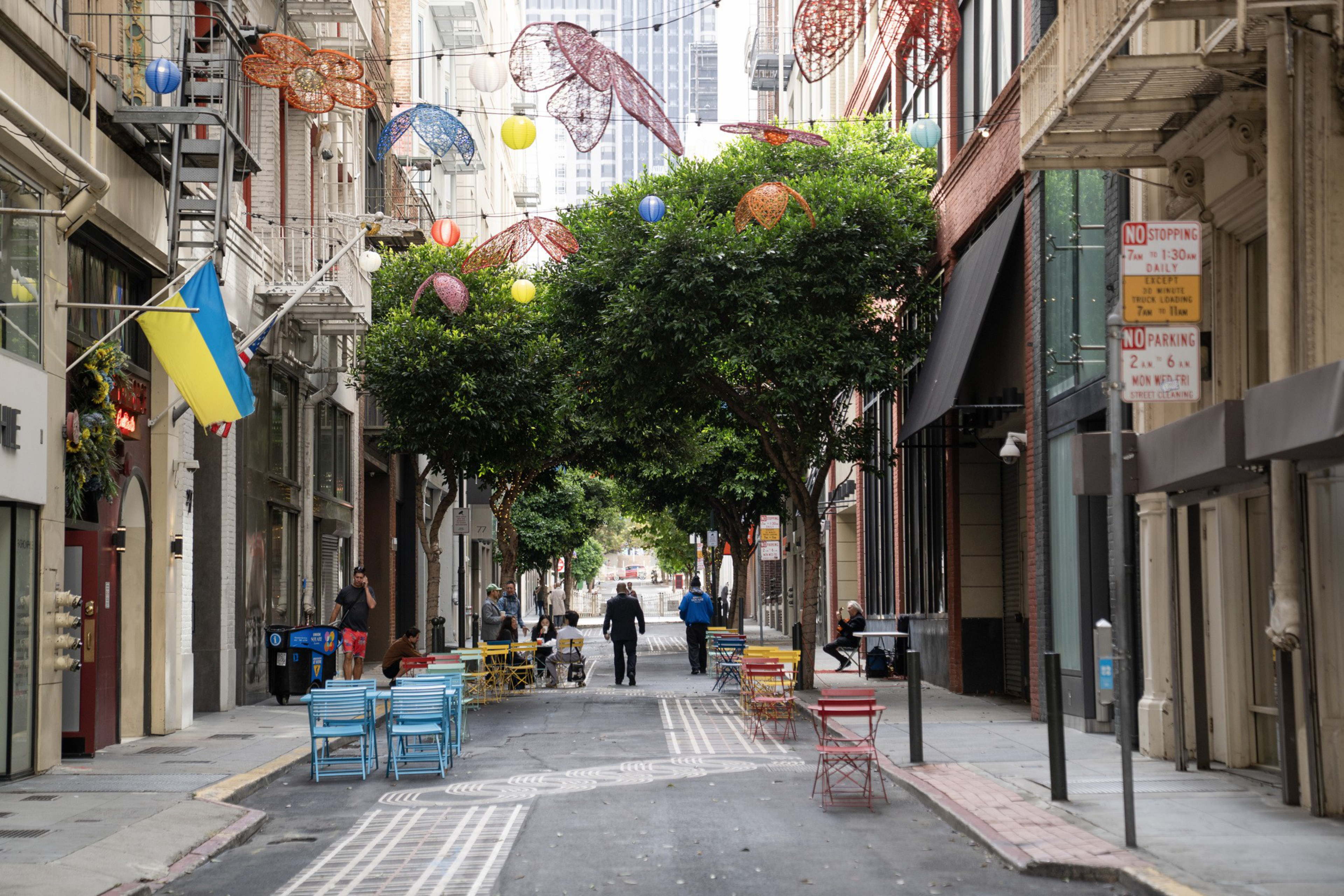 A narrow city street lined with colorful chairs and tables, overhead decorative butterfly lights, green trees, and a Ukrainian flag on the left side.