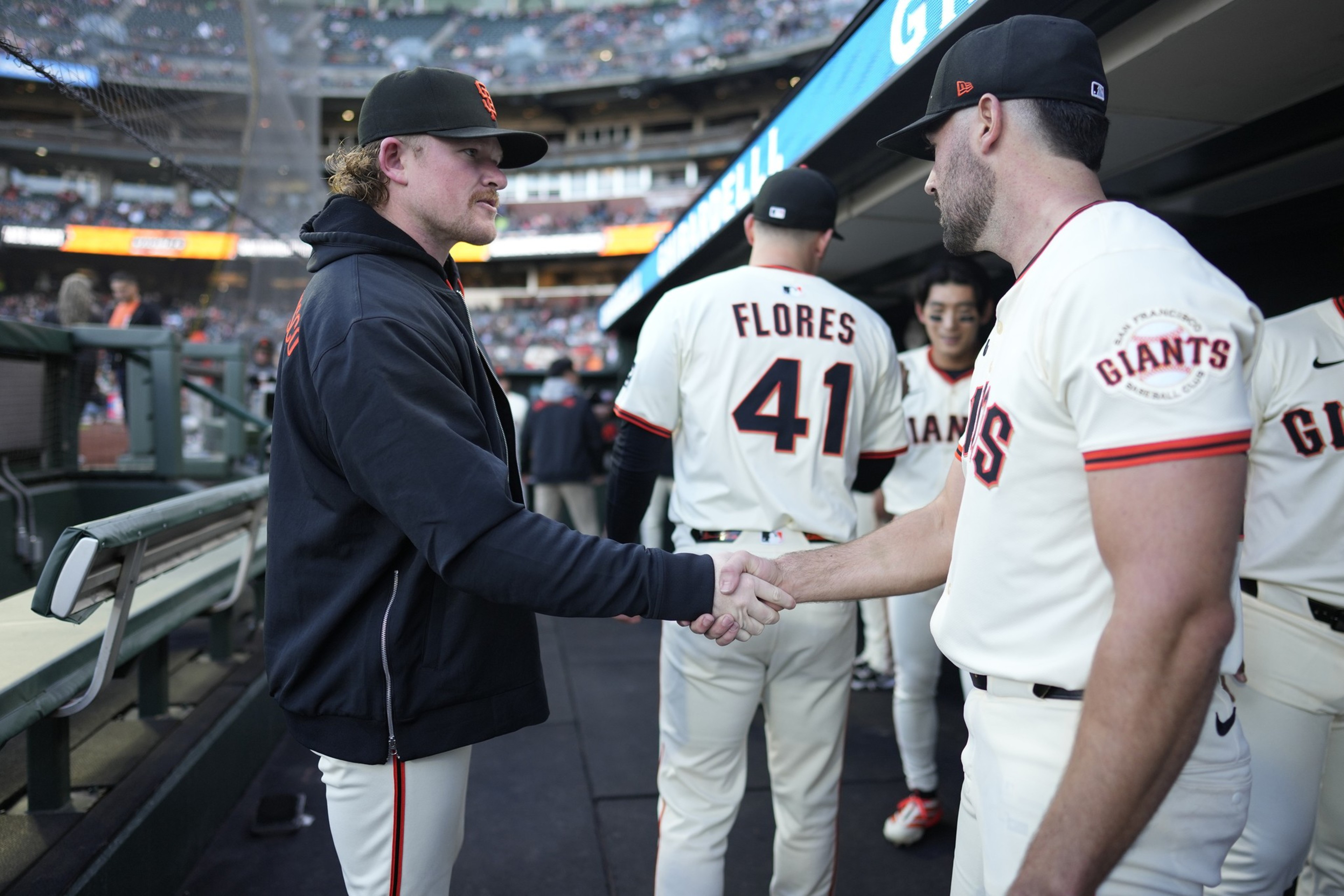 Two men in baseball attire are shaking hands in a dugout. One wears a black jacket, the other, a uniform with "GIANTS" on it. Other players are in the background.