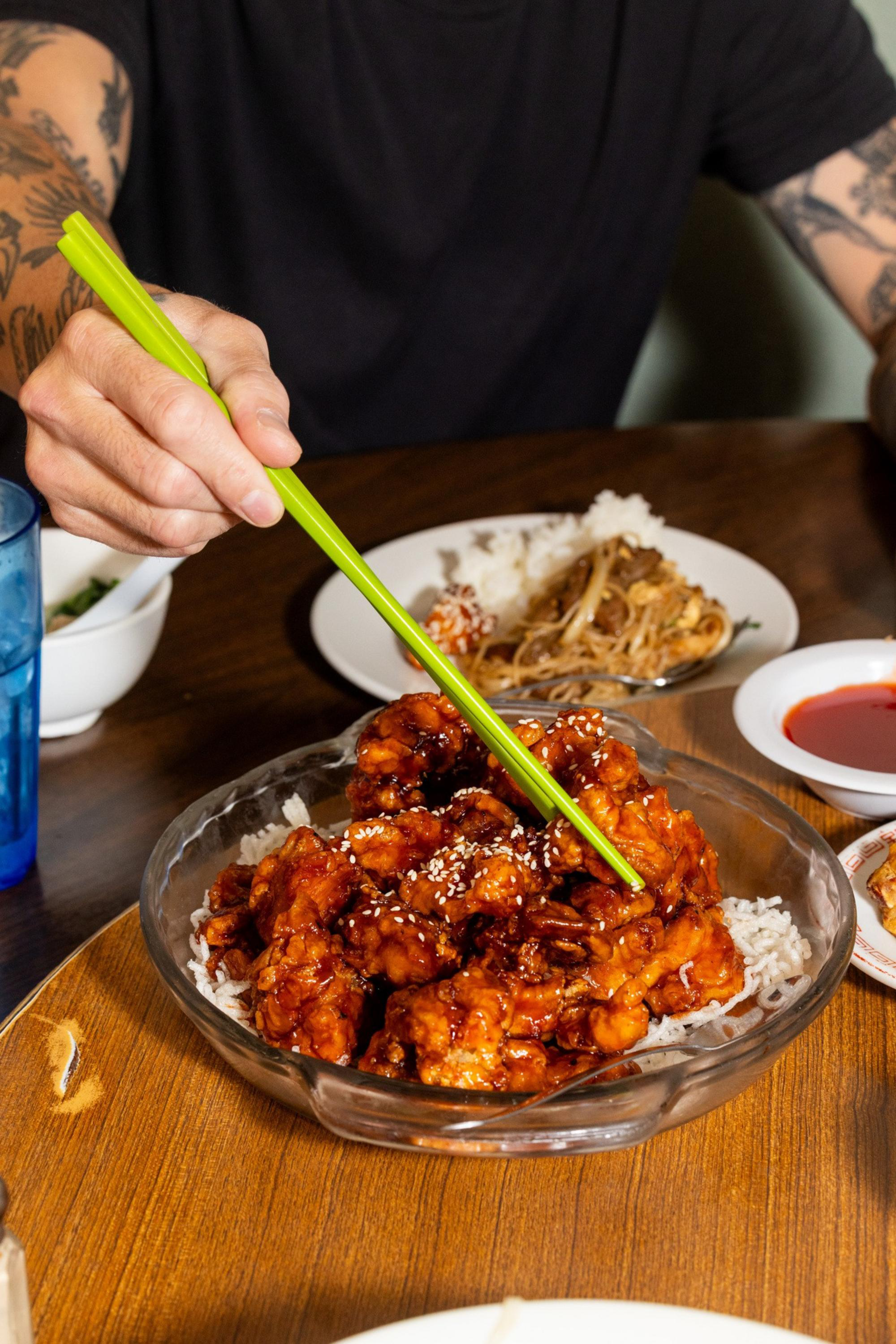 A person with tattoos uses green chopsticks to pick up glazed, sesame-topped chicken pieces from a glass dish with white rice.