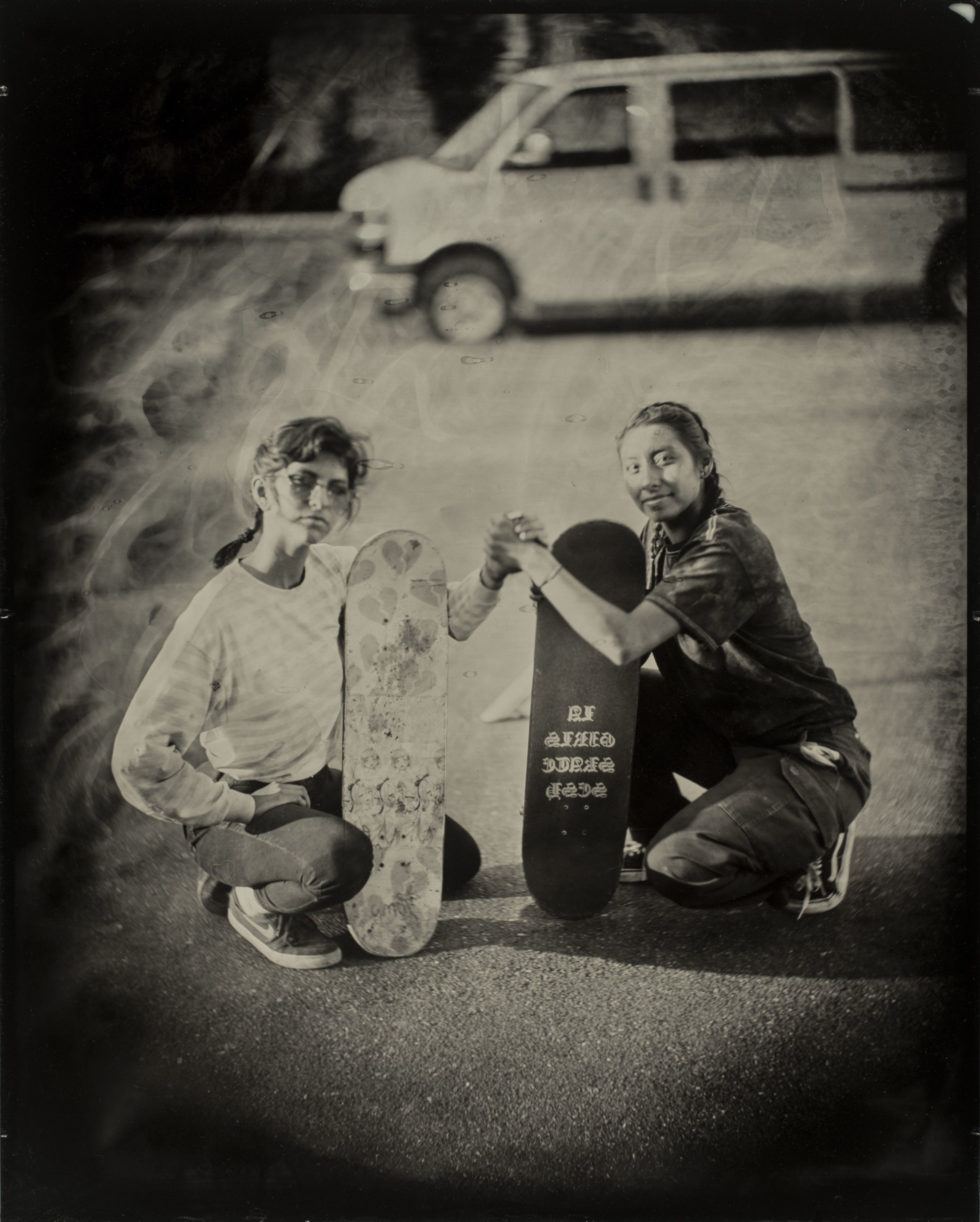 Two young people kneel on pavement, each holding a skateboard. They clasp hands, smiling, with a van in the blurred background. The photo is in a vintage style.