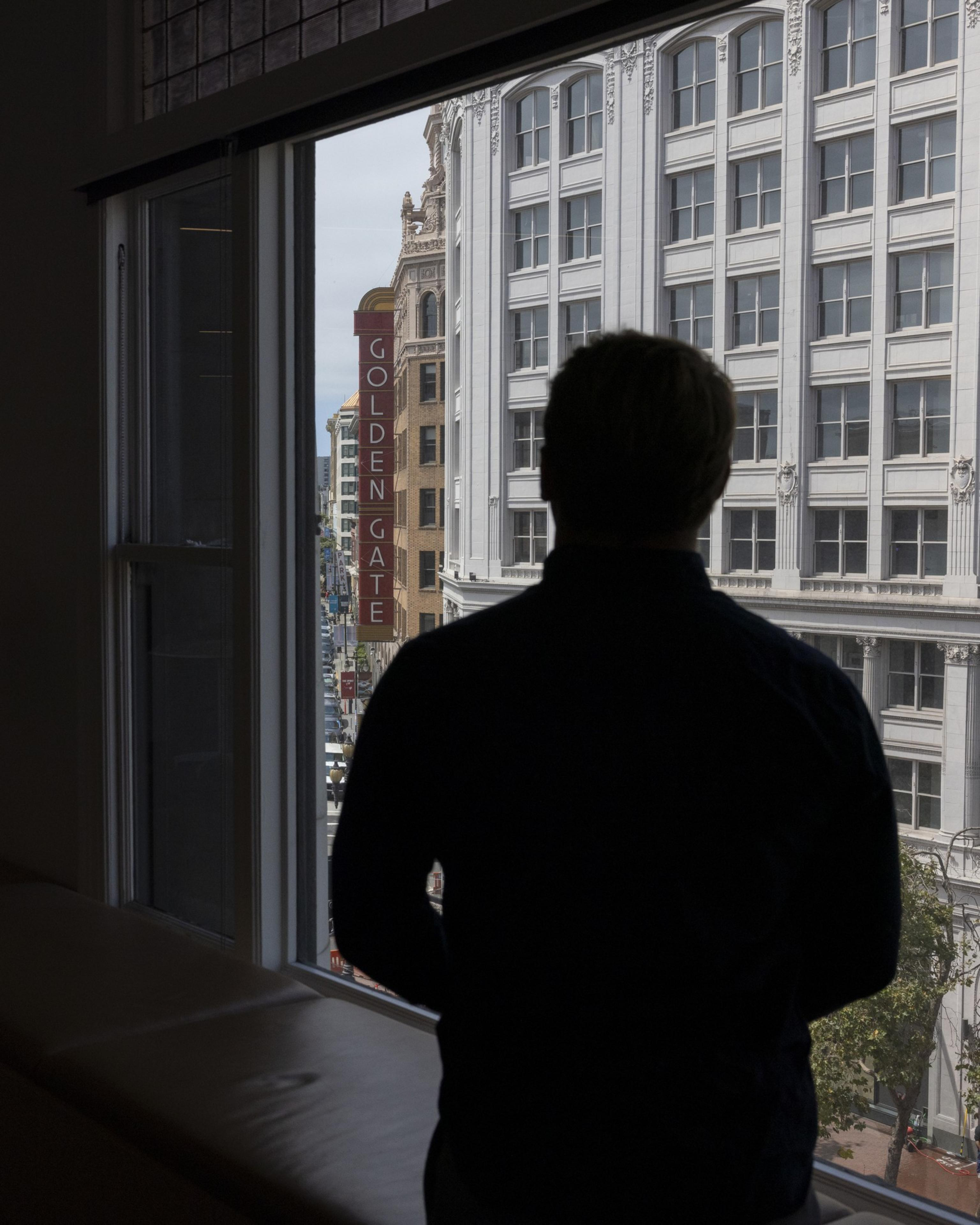 A silhouette of a person stands by a window, looking out at a cityscape with tall buildings. A &quot;Golden Gate&quot; sign is visible on one of the buildings outside.