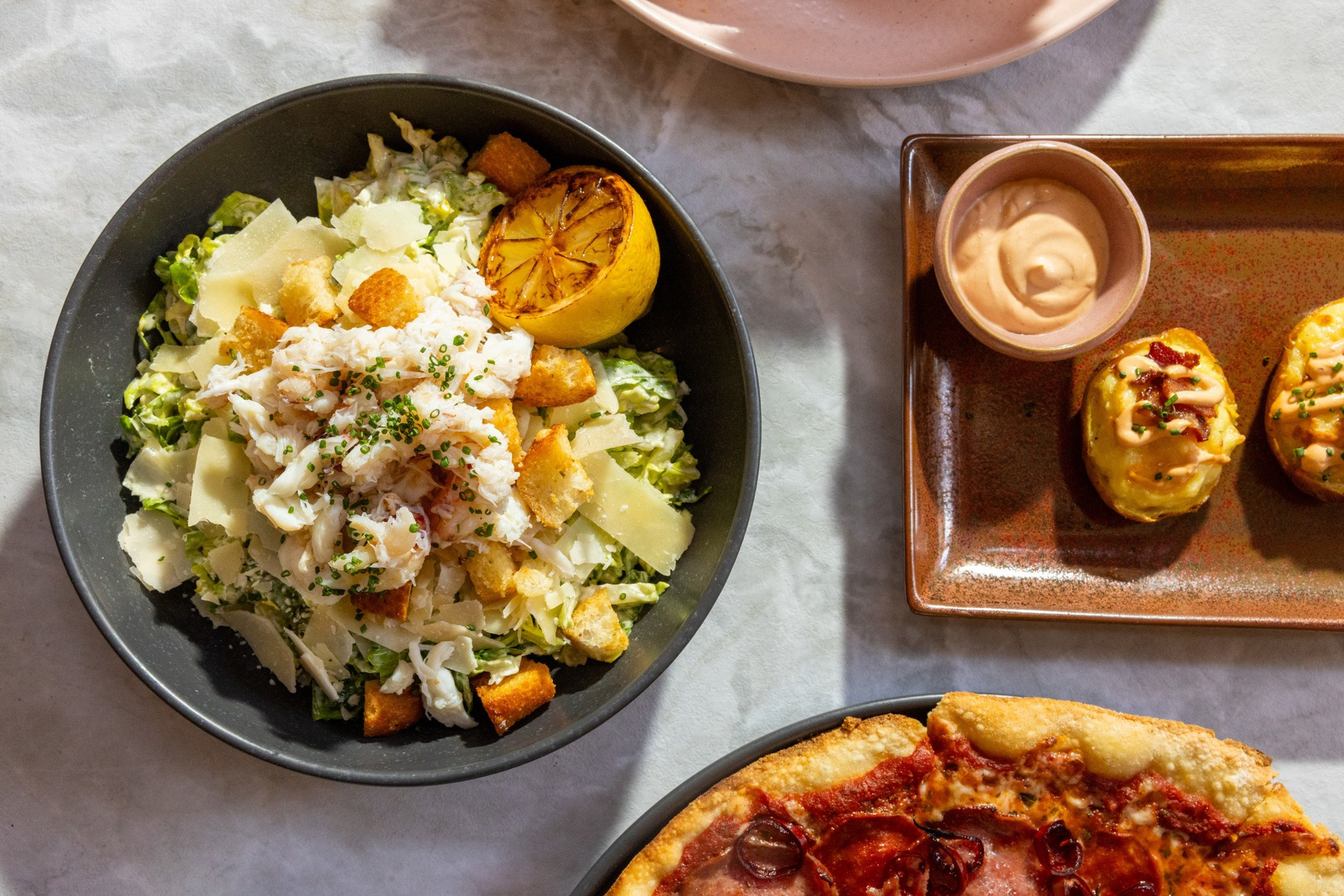 A bowl of Caesar salad with crab meat and grilled lemon, a plate with two stuffed potatoes and dipping sauce, and part of a pepperoni pizza are set on a marble table.