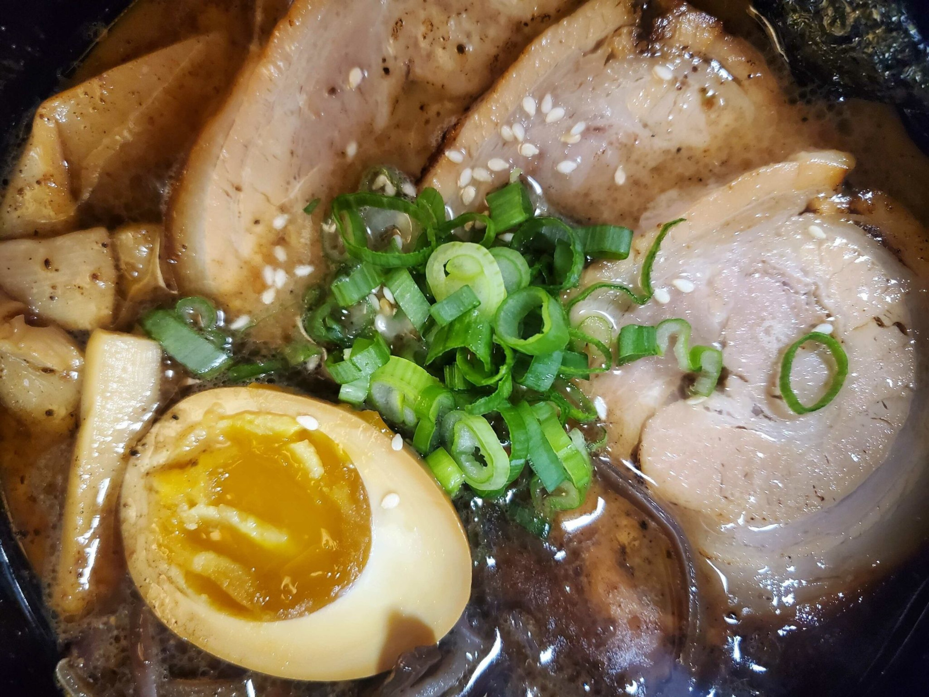 Slices of pork, a halved soft-boiled egg, chopped green onions, and sesame seeds sit atop a rich broth with noodles submerged.
