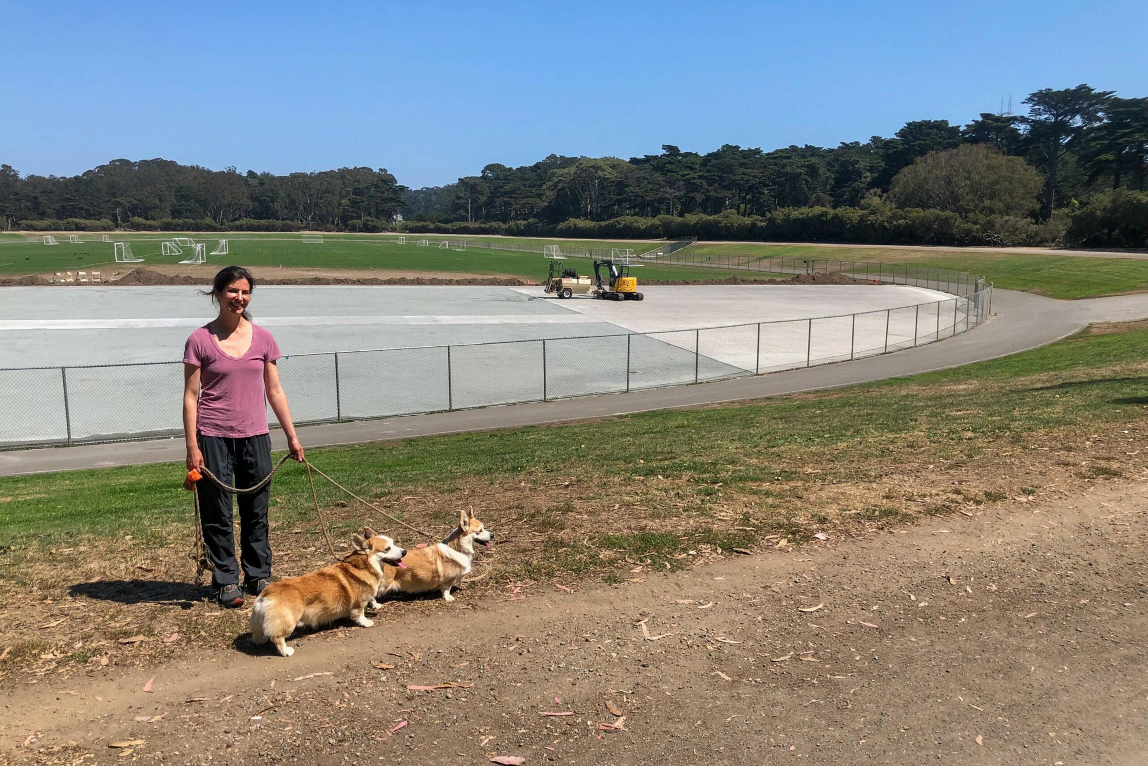 A woman in a purple shirt and black pants stands on a dirt path, holding leashes of two corgis, near a fenced area with a construction vehicle and green fields.