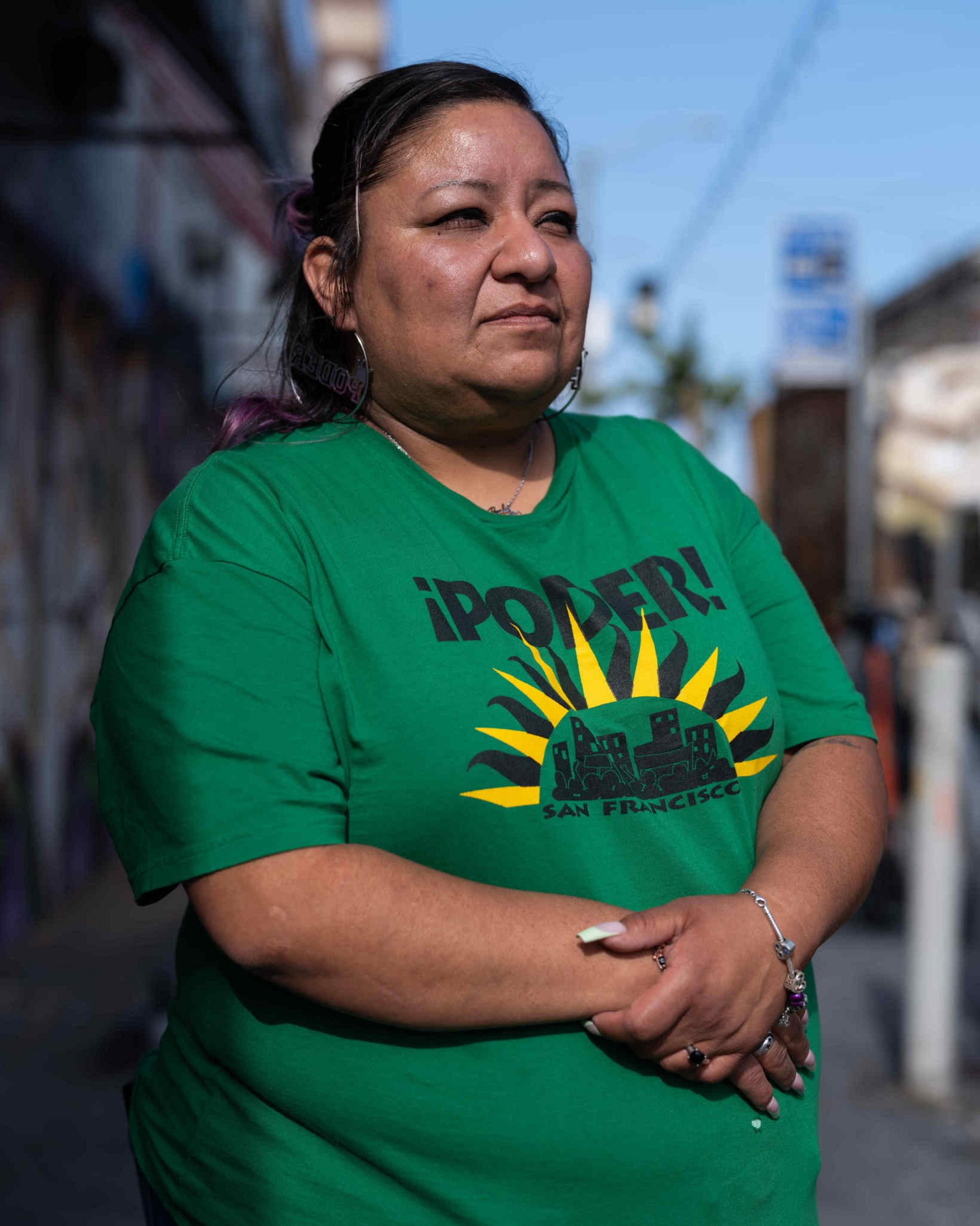 A person with long nails stands outdoors, wearing a green T-shirt with &quot;¡PODER! San Francisco&quot; and a city skyline graphic. They're in an urban setting.