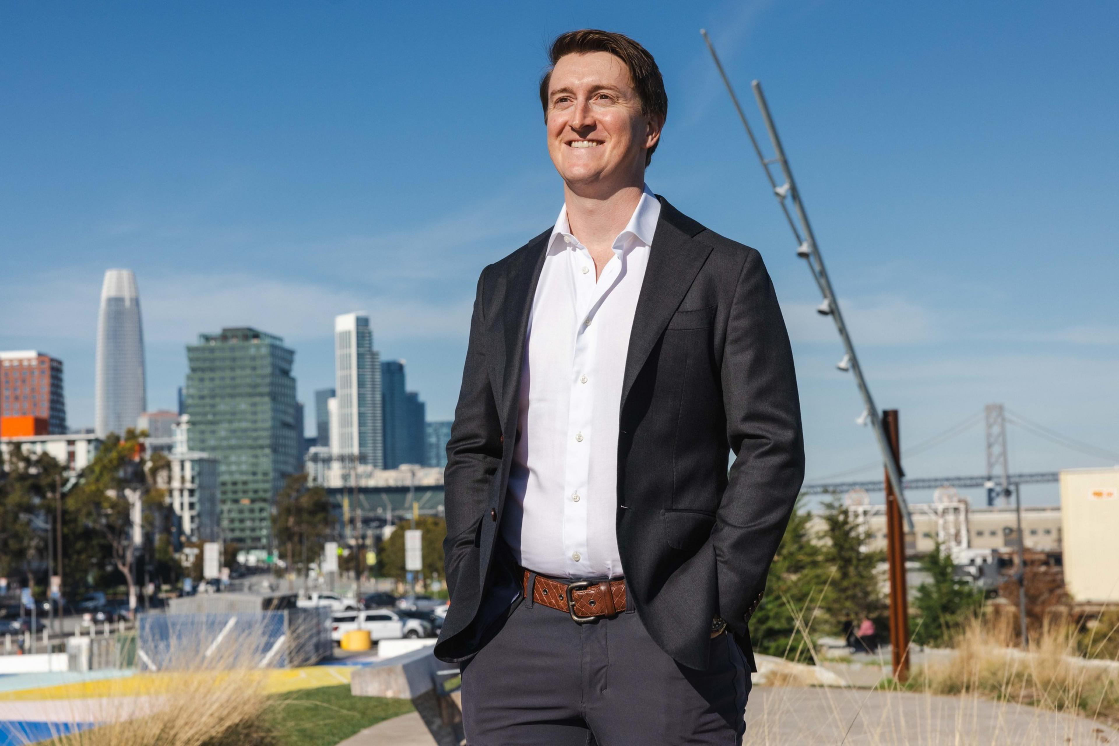 A person in a dark suit and white shirt stands outdoors smiling, with a city skyline and bridge in the background on a clear day.