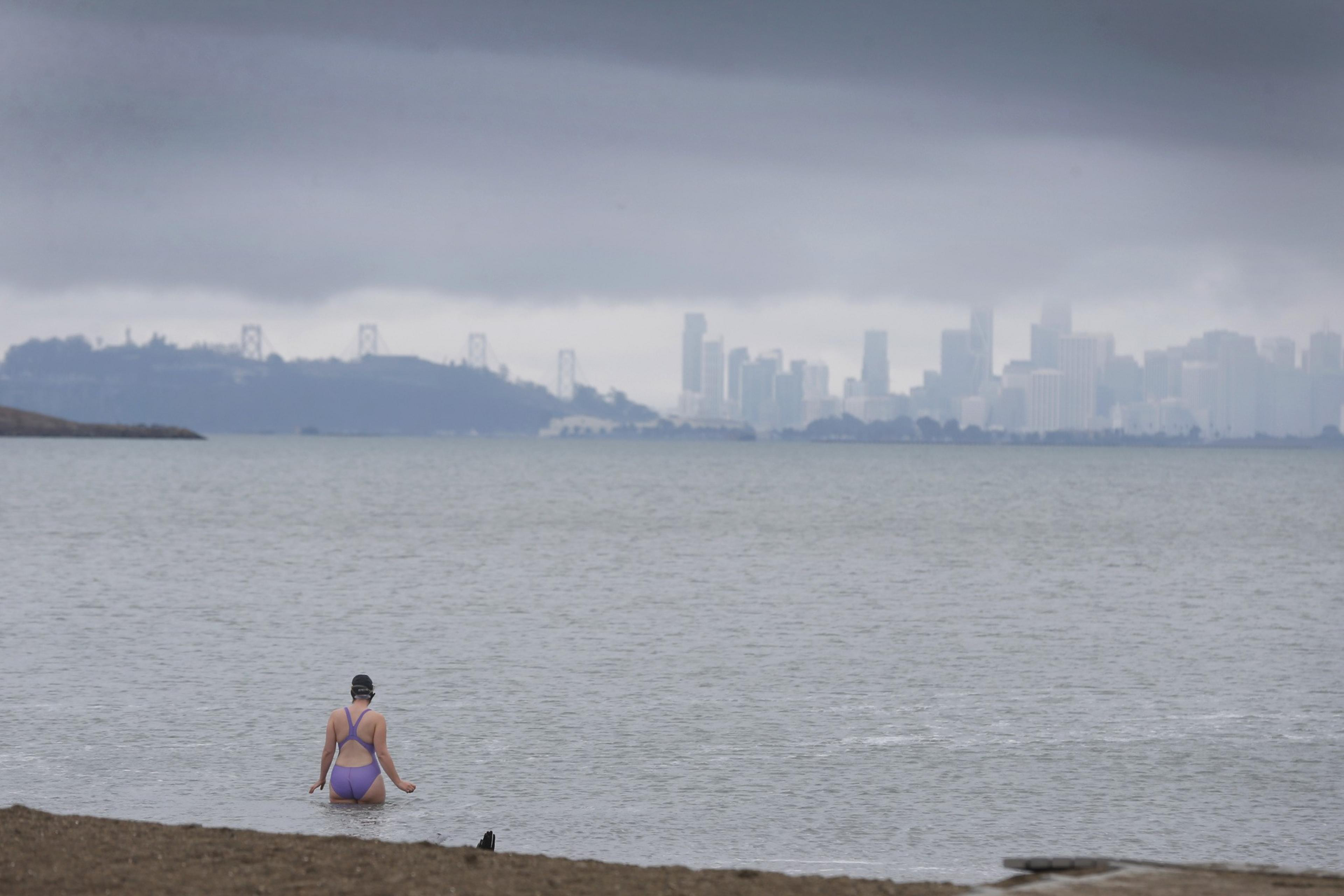A person in a swimsuit sits by the water, looking at a foggy city skyline in the distance.