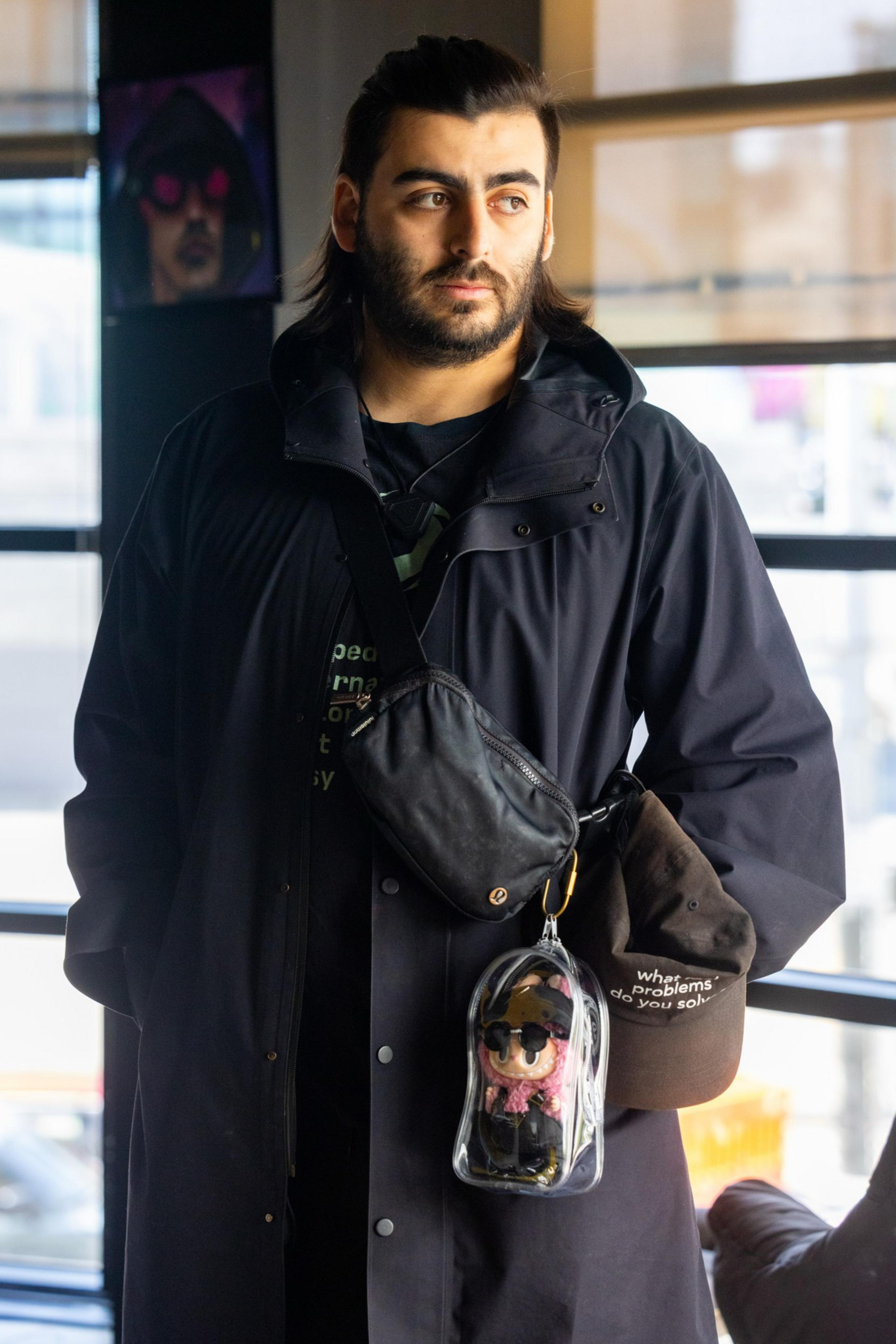 Sure! A person with long dark hair is wearing a black coat and crossbody bag. They're holding a hat and a clear pouch containing a figure.