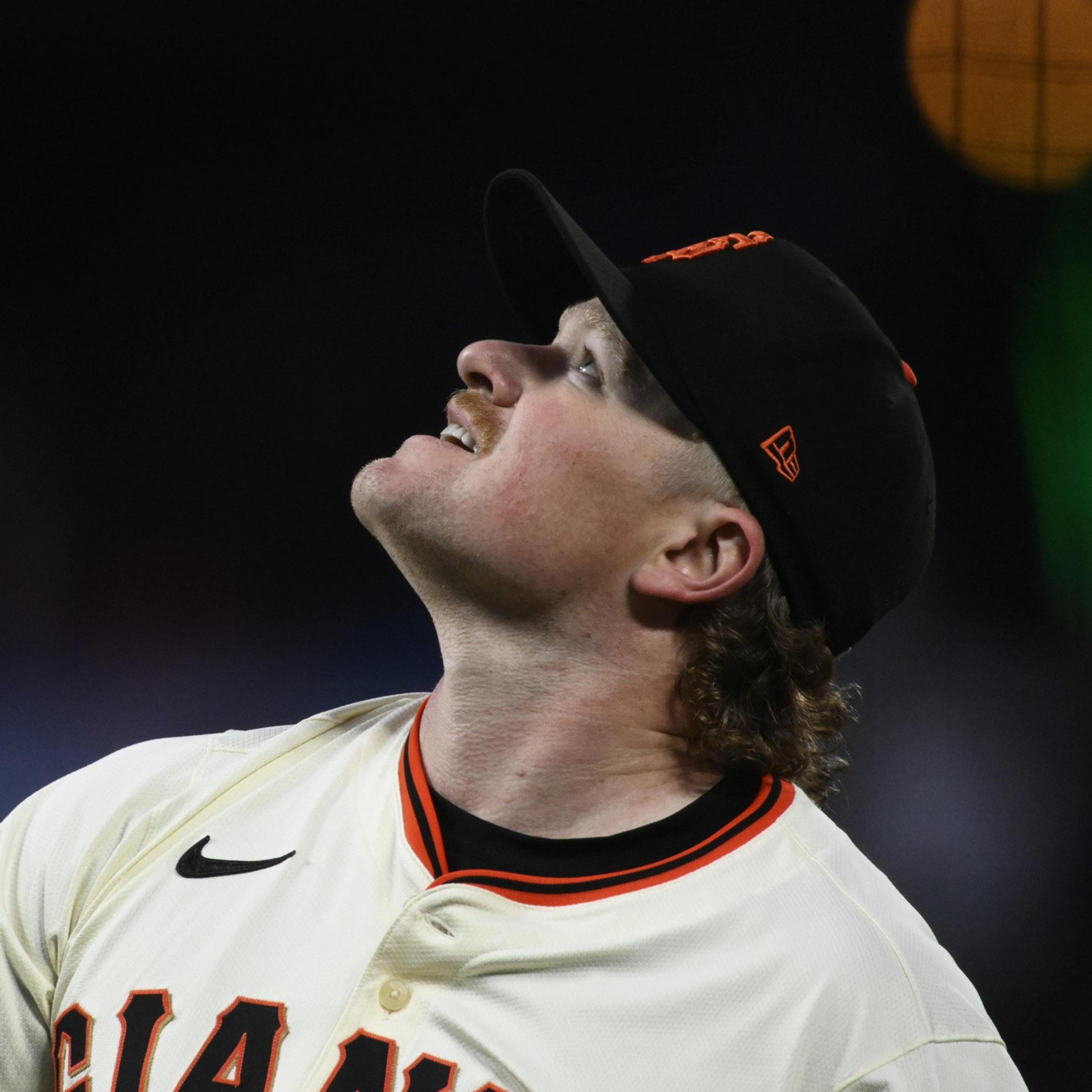A baseball player in a white uniform with orange and black details looks upwards. He wears a black cap, and the background is dark with blurred lights.