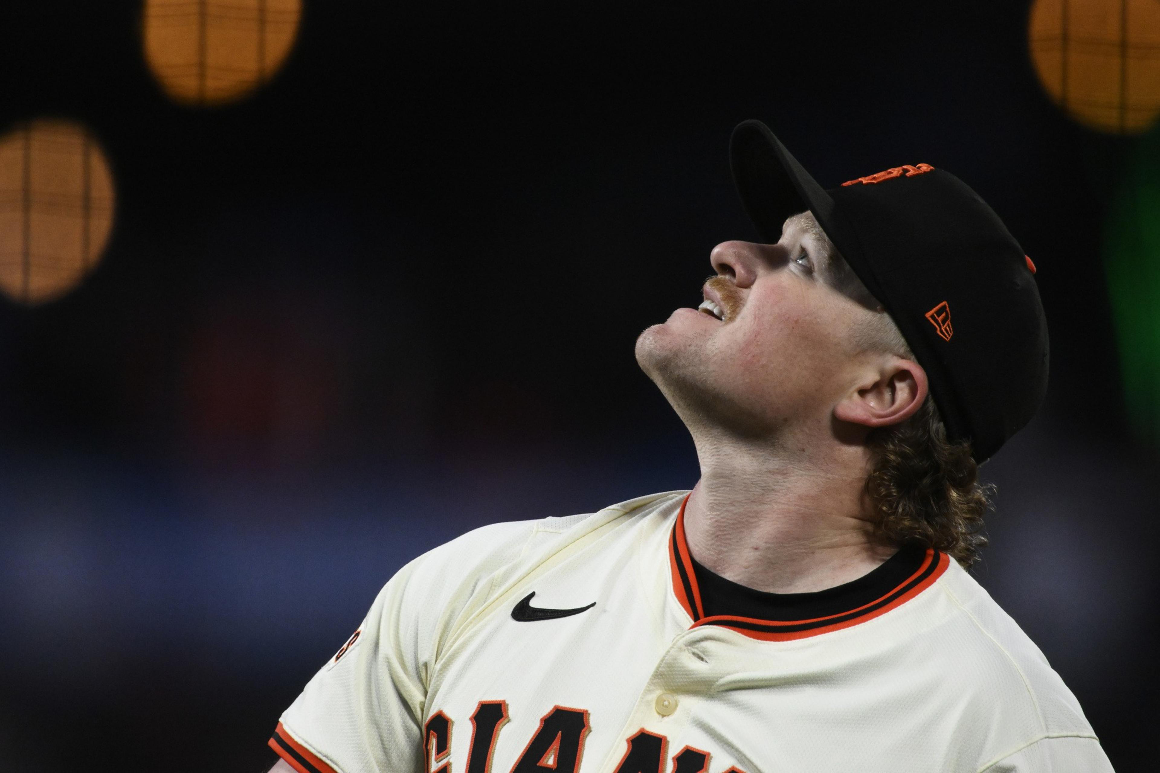 A baseball player in a white uniform with orange and black details looks upwards. He wears a black cap, and the background is dark with blurred lights.