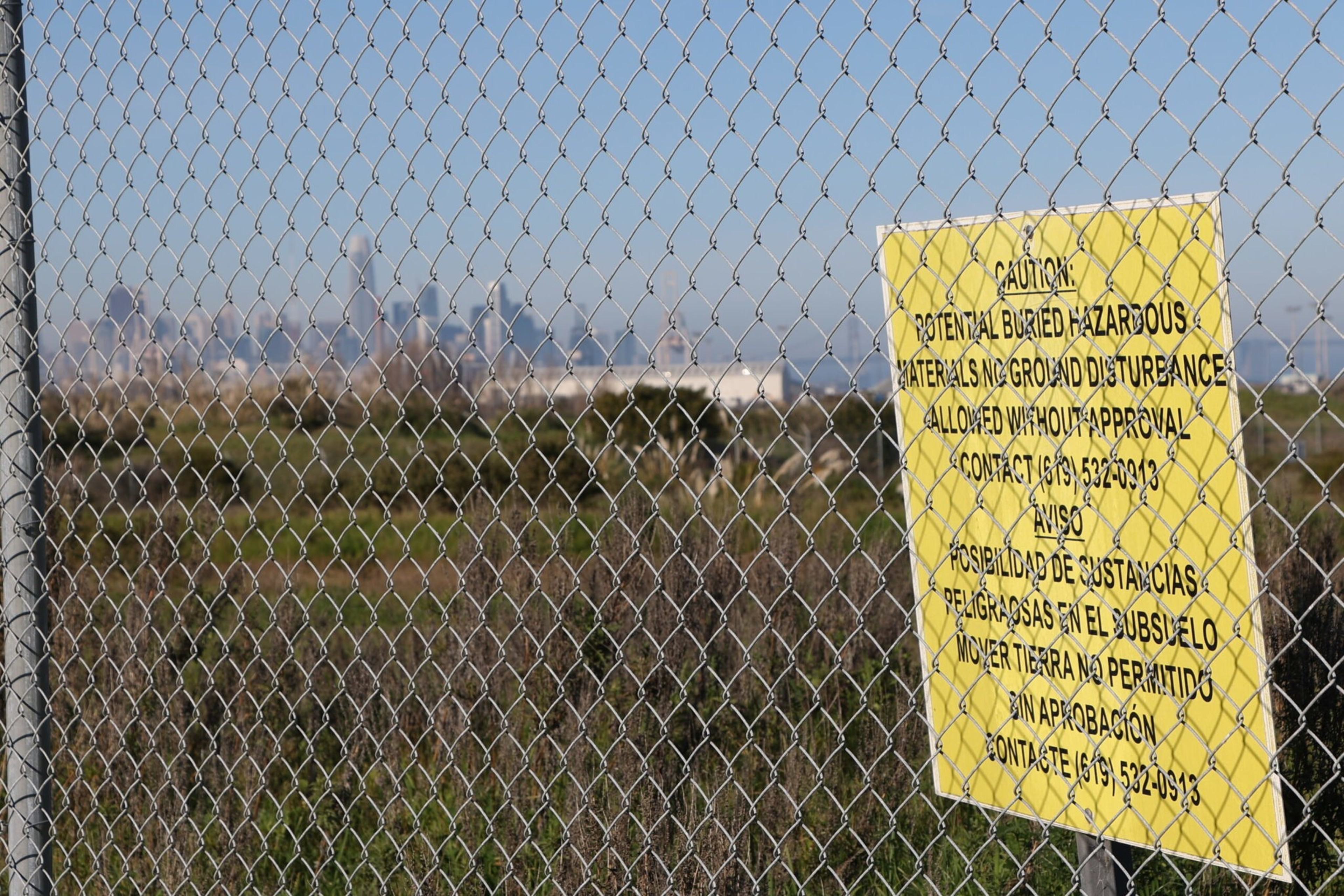 A chain-link fence with a yellow caution sign warns of potential buried hazardous materials and restricts ground disturbance without approval.