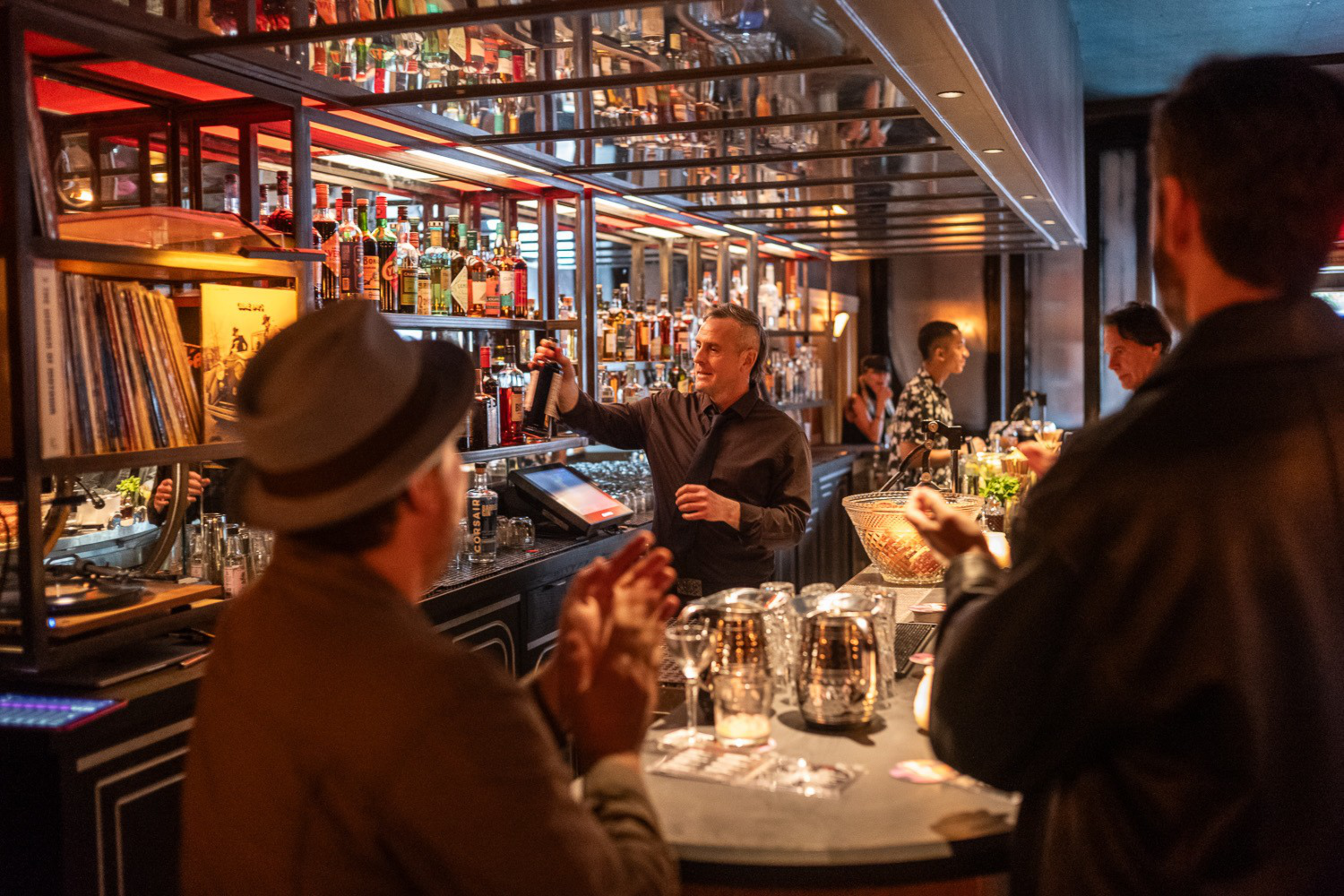 A bartender pours a drink behind a stocked bar while two patrons, one wearing a hat, watch and clap in a warmly lit bar setting.