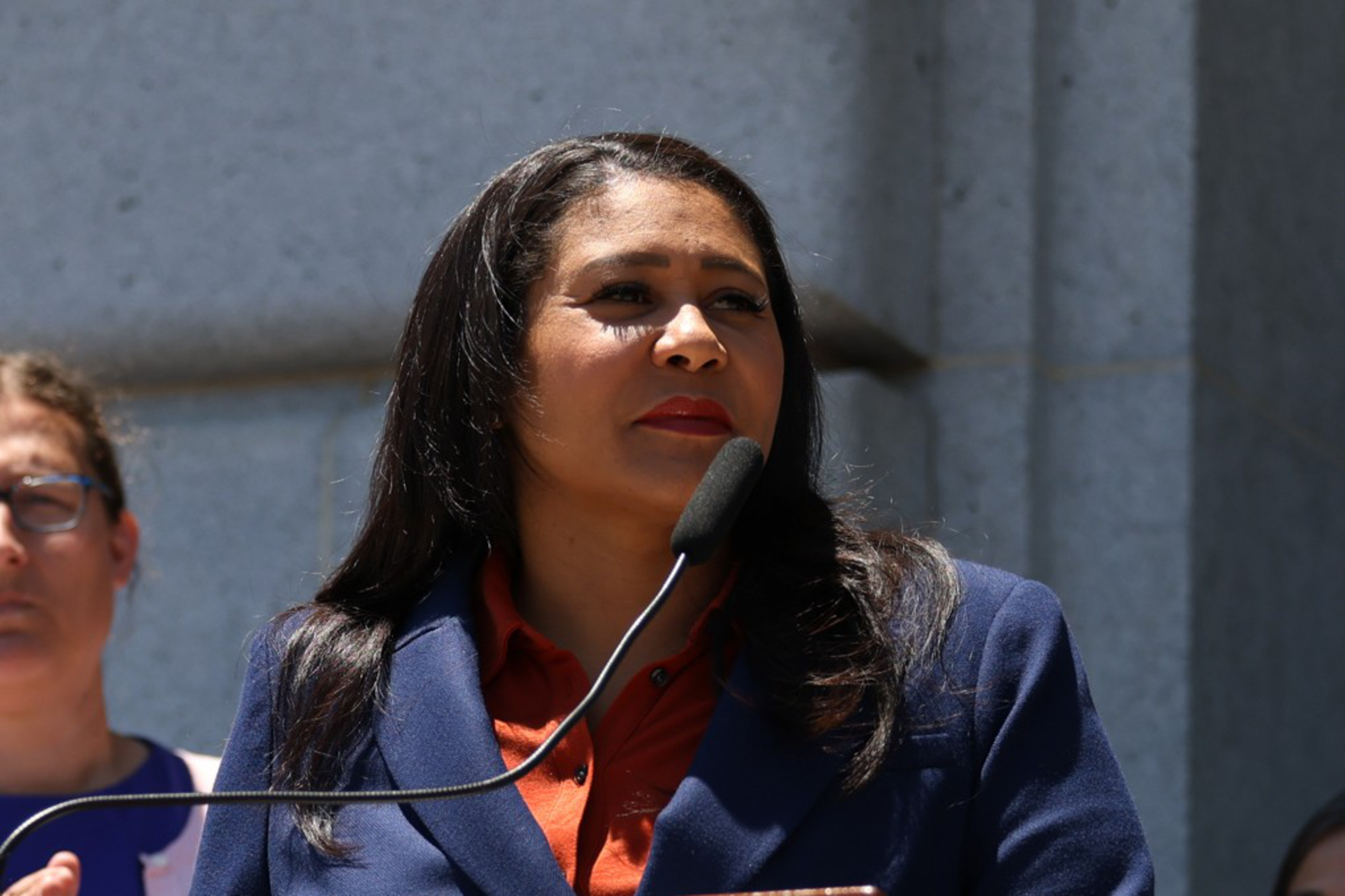 A woman in a navy blazer and red shirt speaks into a microphone, standing outdoors with a gray stone wall behind her and a person partially visible nearby.
