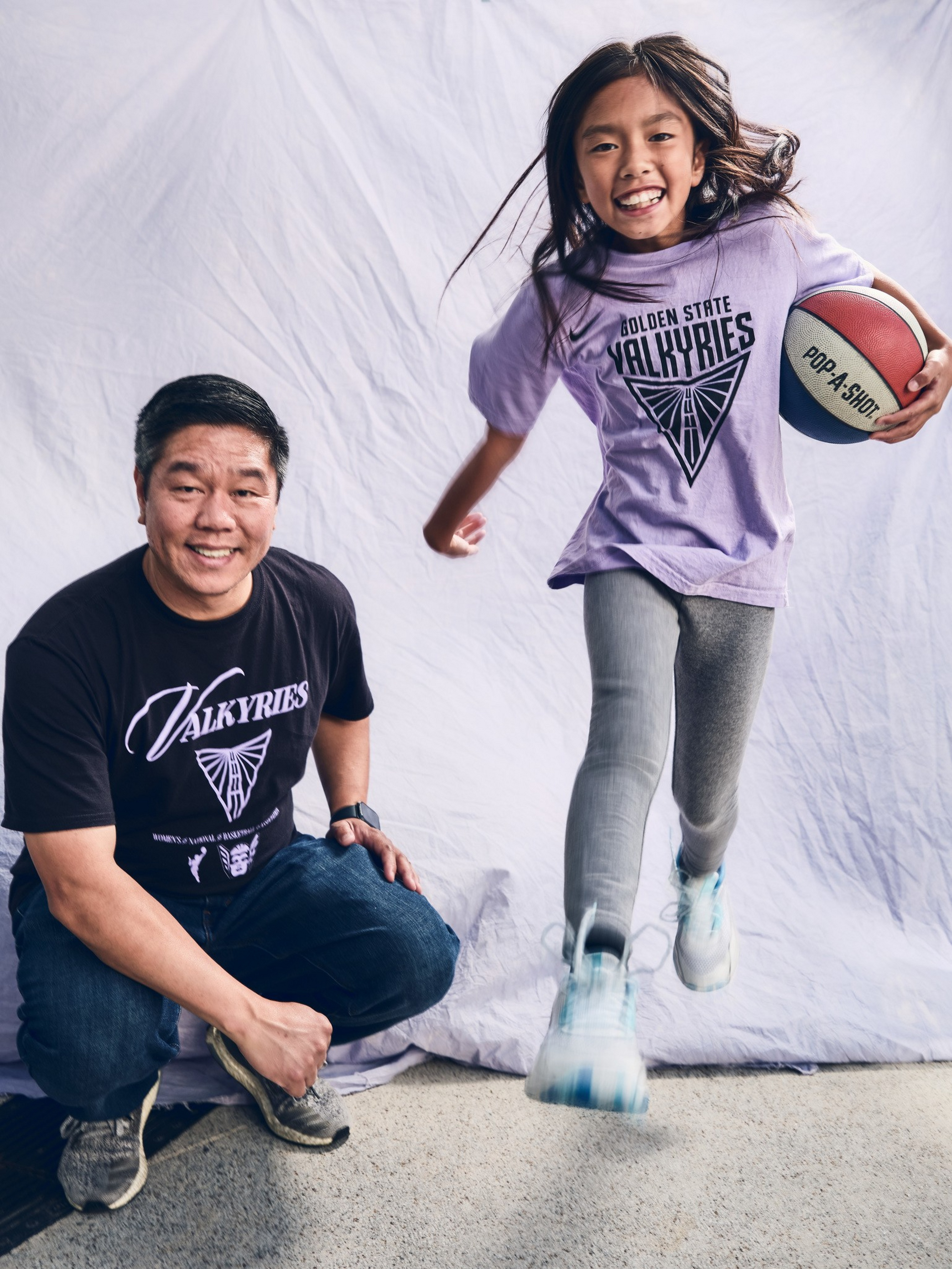 A smiling man crouches wearing a black “Valkyries” shirt, while a girl in a purple “Golden State Valkyries” shirt runs holding a red, white, and blue basketball.