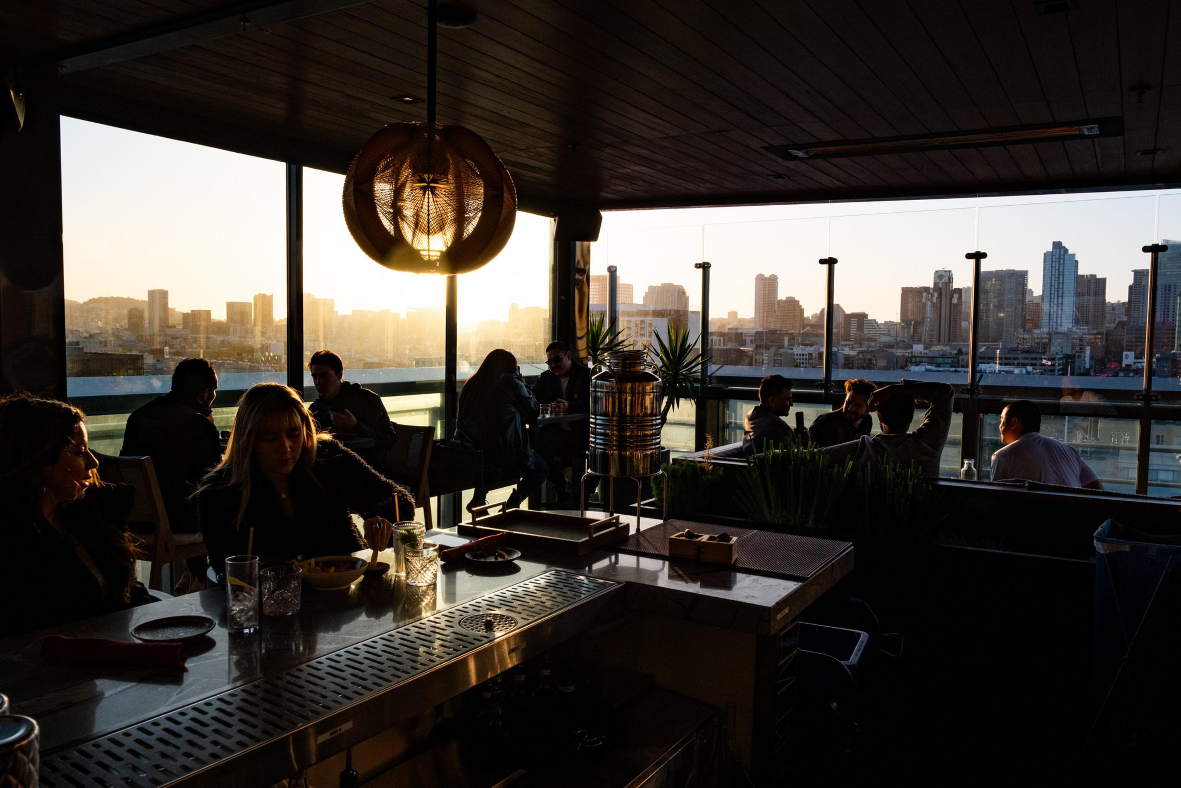 A rooftop bar during sunset, with people sitting at tables and enjoying drinks. The skyline of a city is visible through large windows.
