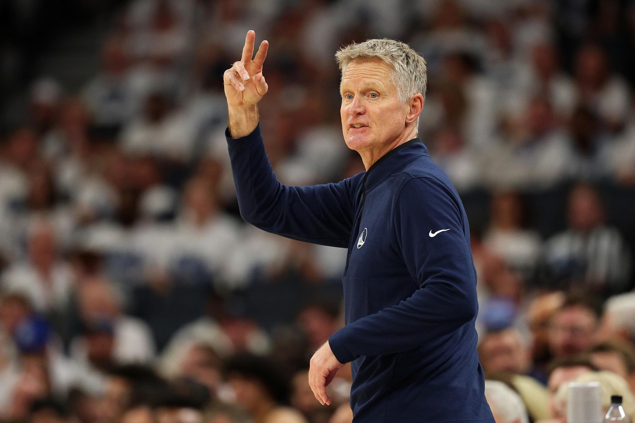A man with short gray hair wearing a navy blue long-sleeve shirt raises his hand, making a peace sign, with a blurred crowd in the background.