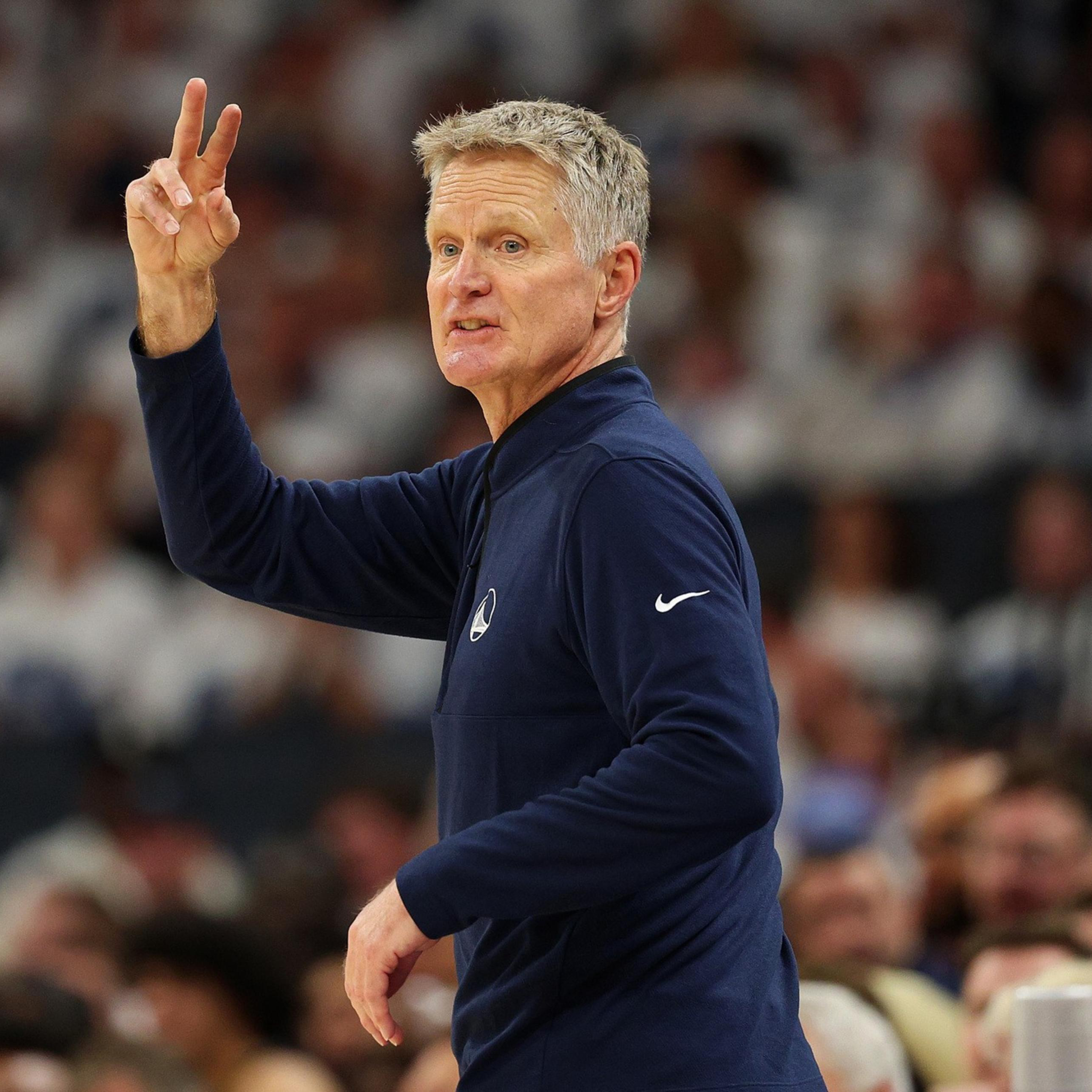 A man with short gray hair wearing a navy blue long-sleeve shirt raises his hand, making a peace sign, with a blurred crowd in the background.