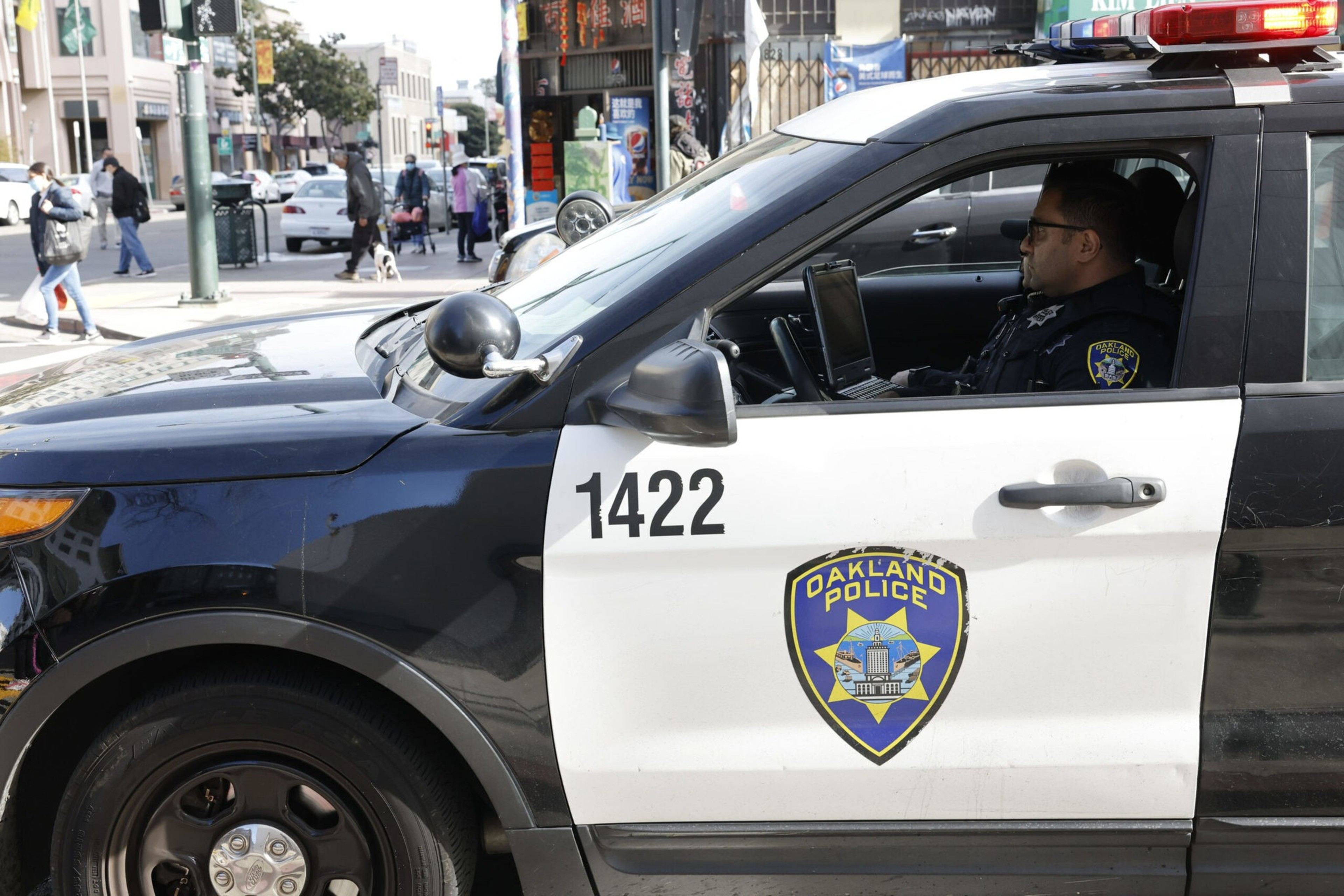 A police officer in an Oakland Police vehicle labeled "1422" is parked on a busy city street, working on a laptop. Pedestrians and cars are visible in the background.
