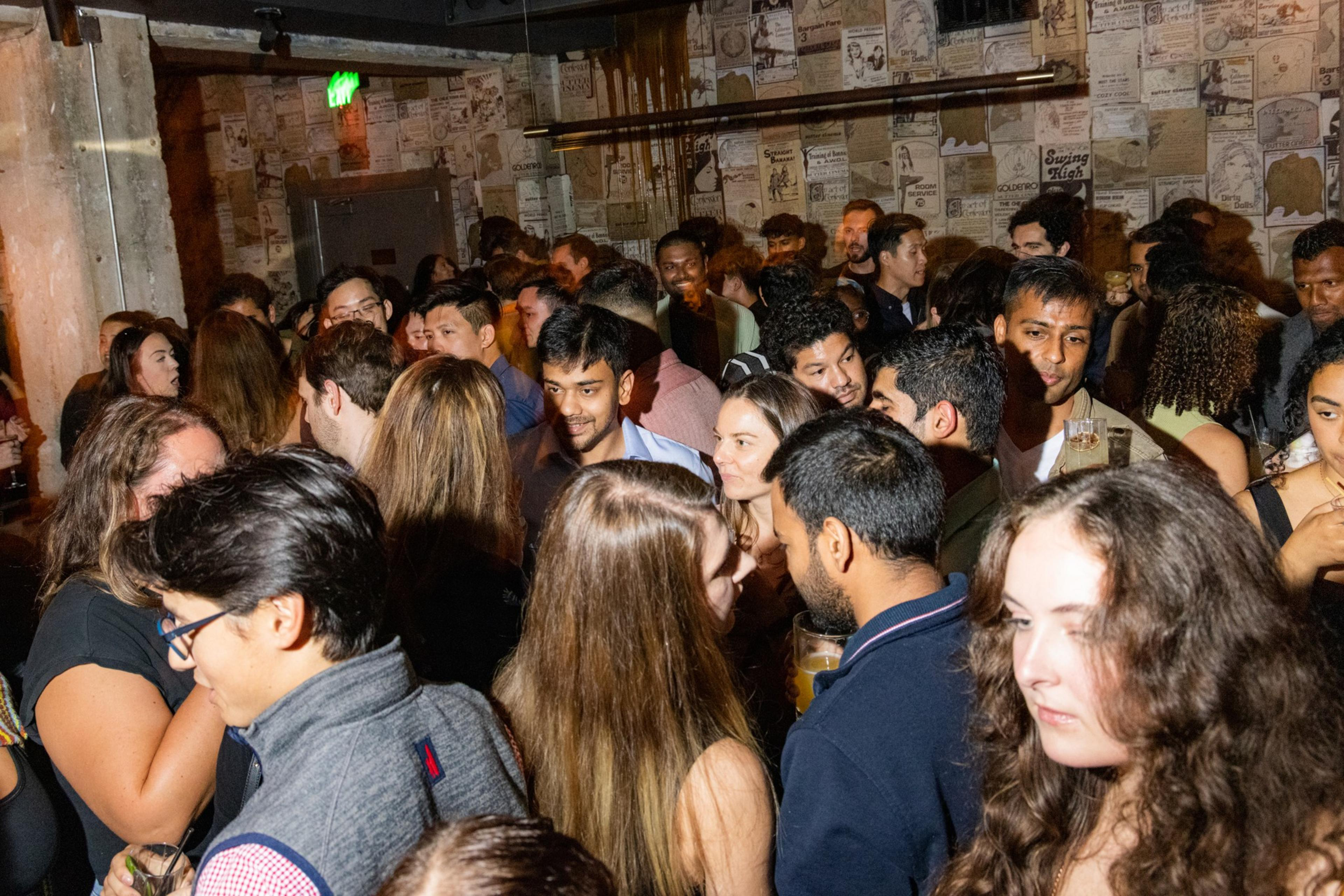 A crowded indoor gathering shows people mingling and conversing. The walls are covered in vintage posters, and some attendees are holding drinks, creating a lively atmosphere.