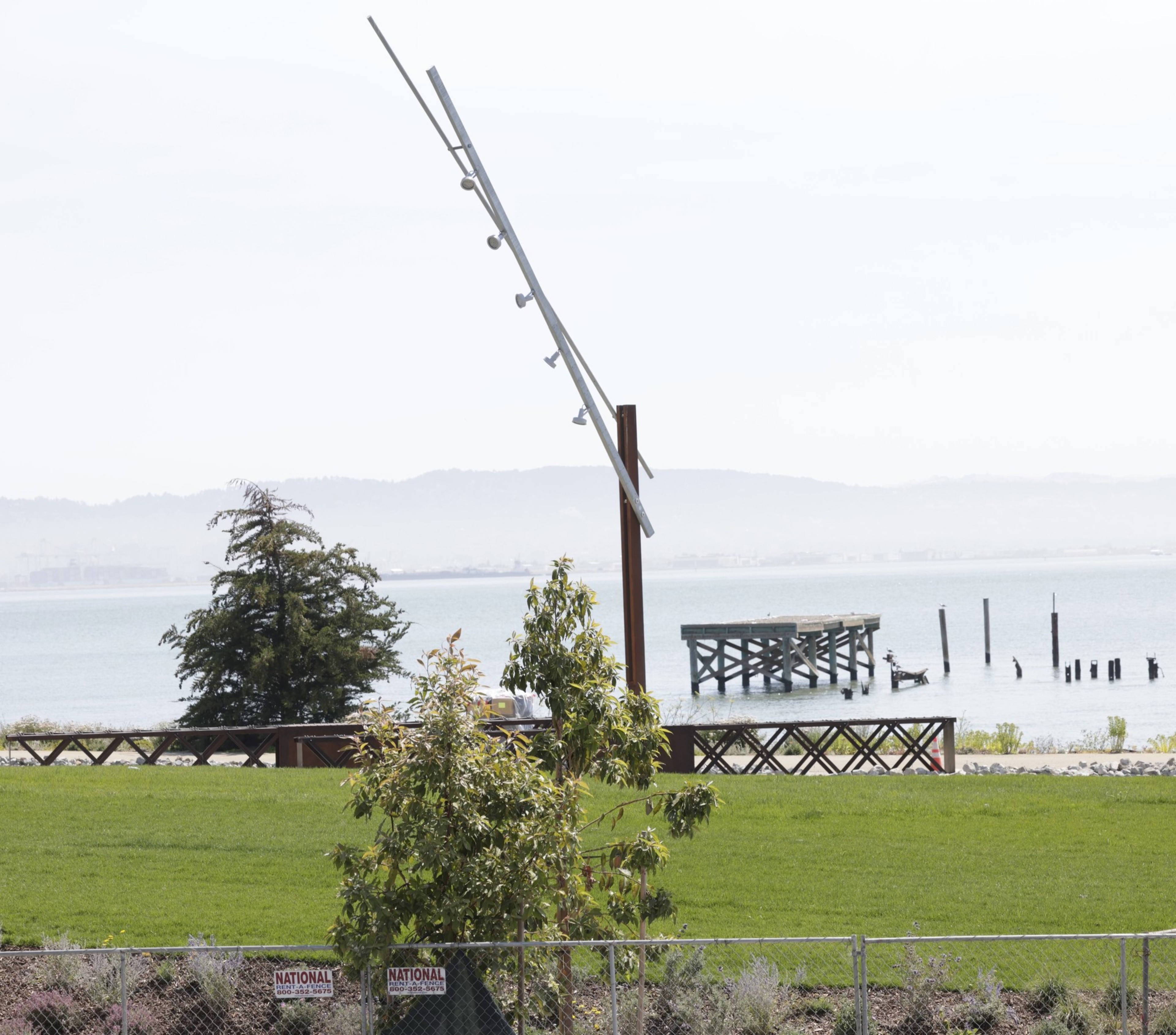 The image shows a grassy area with some trees near a body of water. There's a modern, angled light pole and a wooden pier extending into the water. A mountain range is in the background.