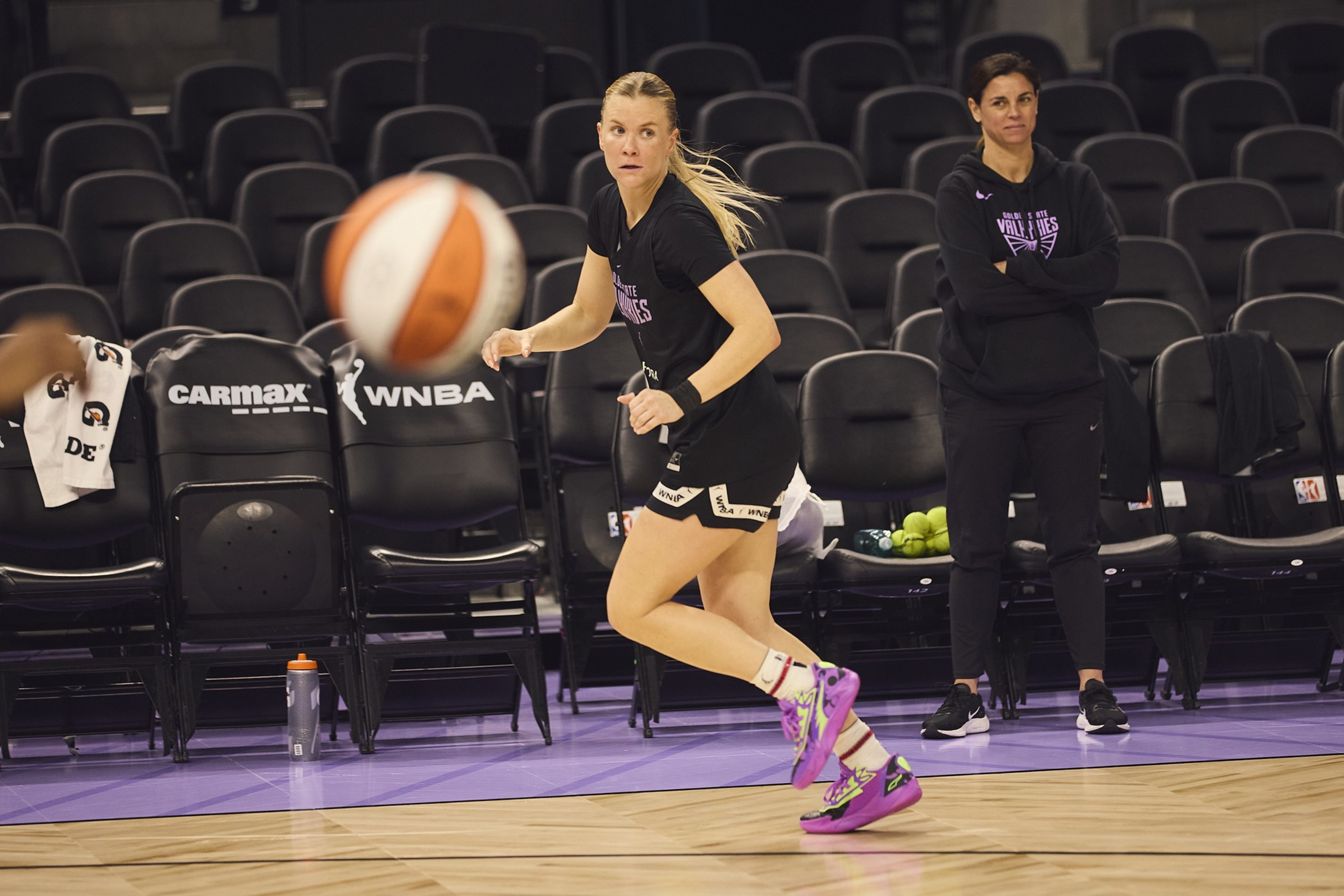 A woman in athletic gear is mid-jump with a basketball nearby. Another person watches from the sidelines. Empty chairs and sports equipment are visible.