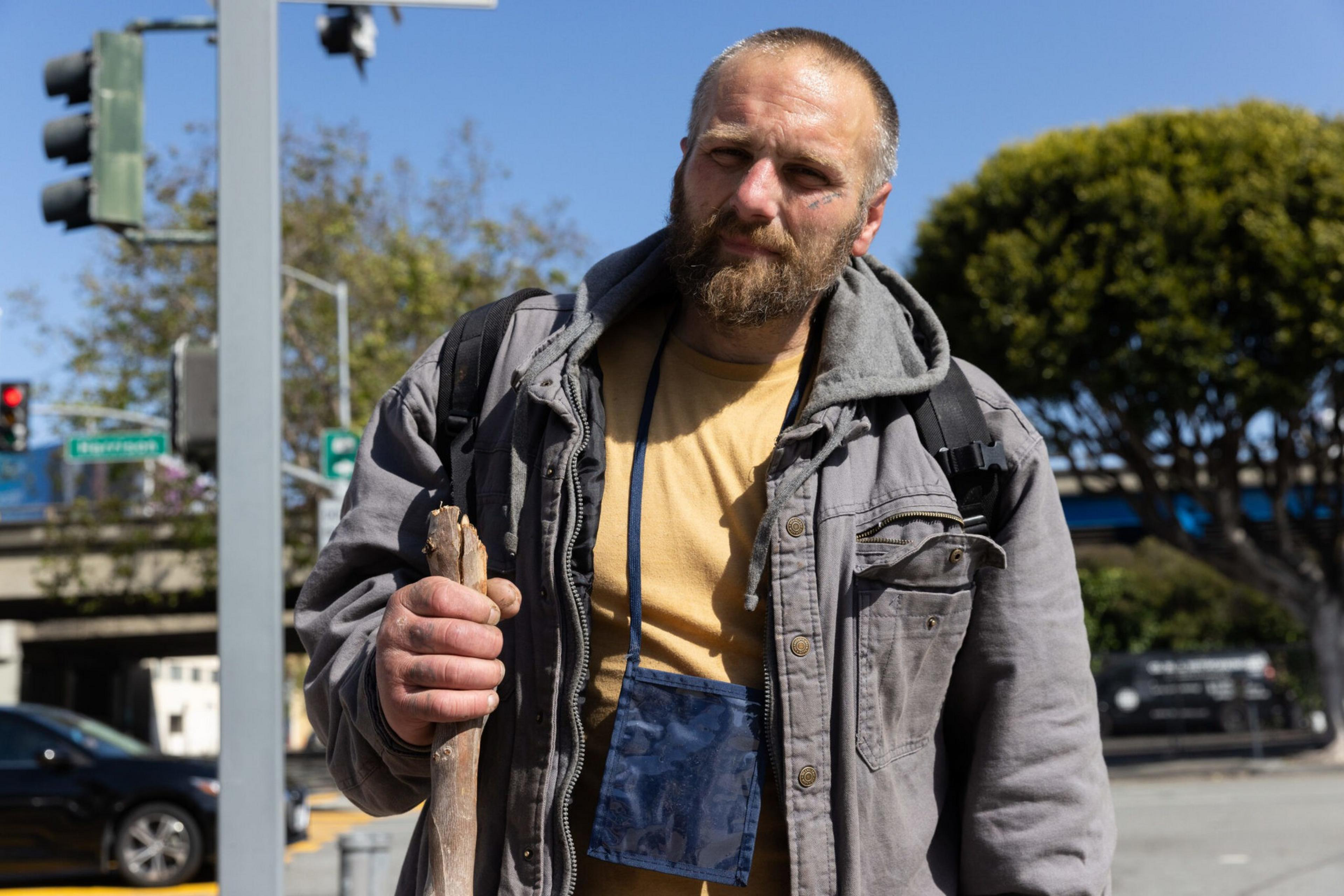 A bearded man in a gray jacket and yellow shirt holds a wooden stick while standing outdoors near a street intersection.