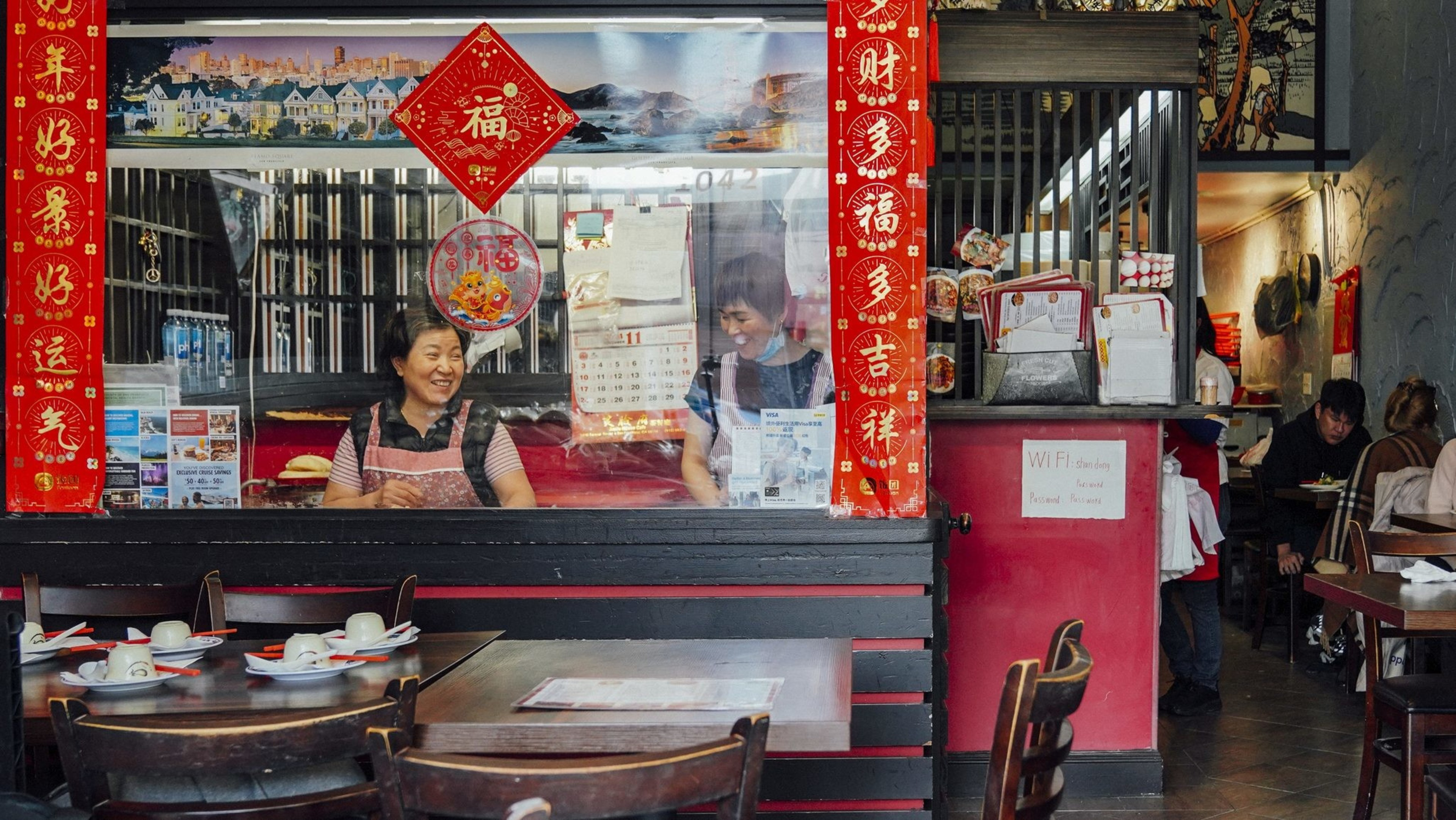 The image shows a cozy Chinese restaurant with red decorations. Two people stand behind a counter smiling, and tables with cups and saucers are in the foreground.