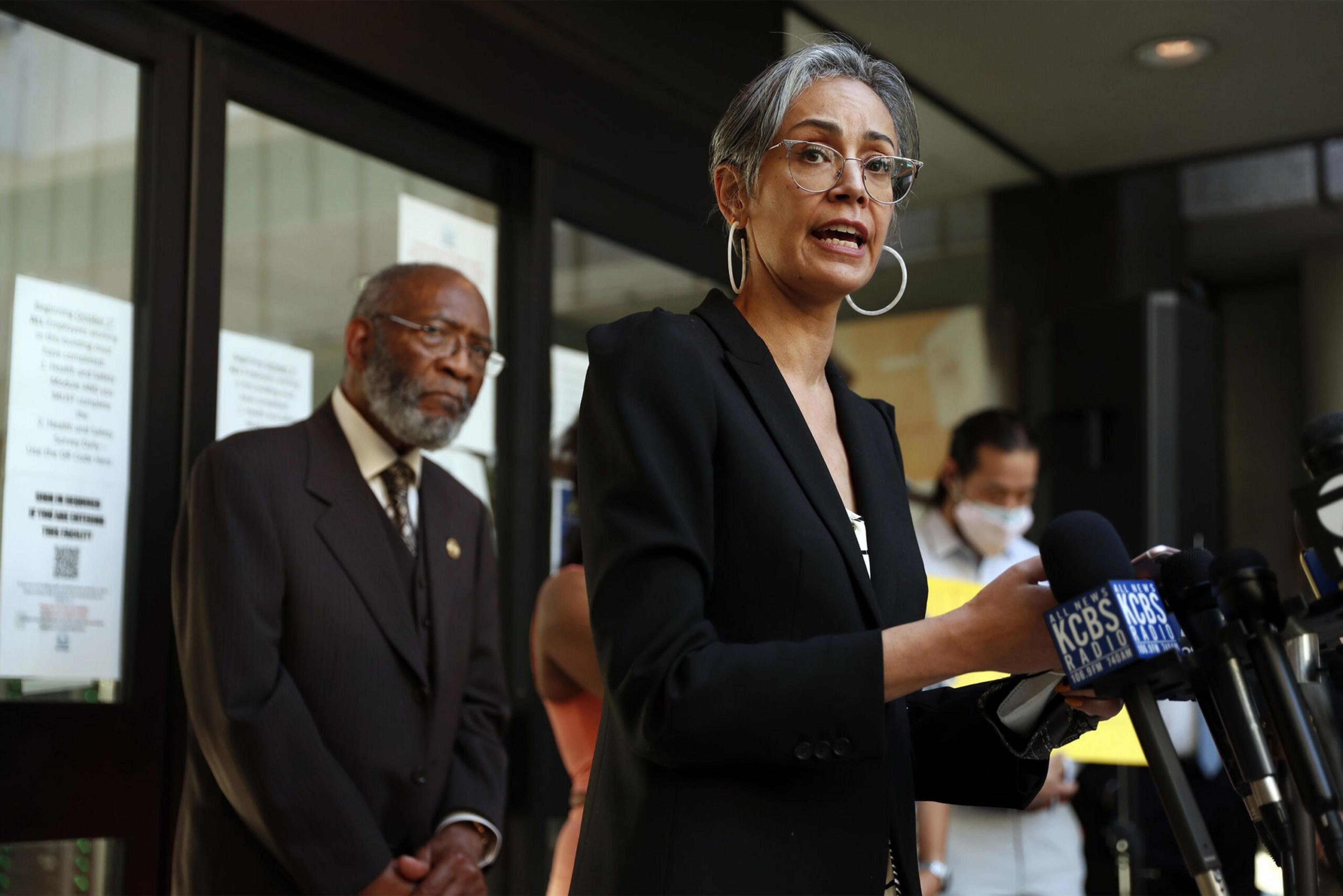 A woman in glasses and a black blazer speaks at a podium with multiple microphones, while two people stand behind her outside a building.
