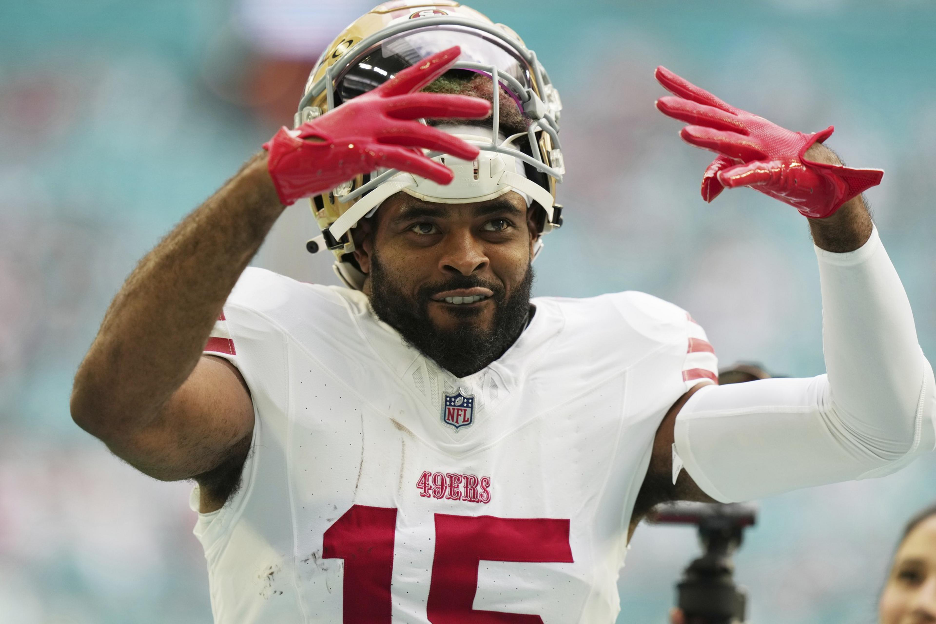 A football player in a San Francisco 49ers uniform with number 15 raises both hands wearing red gloves, with his helmet pushed up on his head.