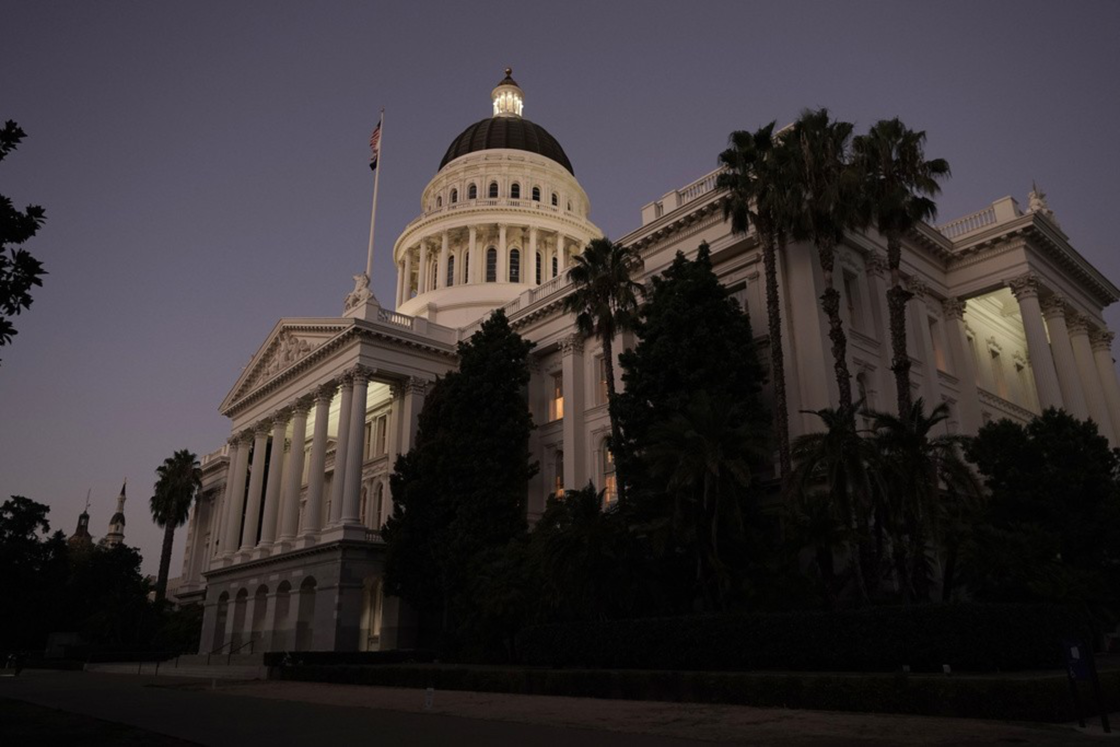 A neoclassical building with a lighted dome at dusk, surrounded by palm trees under a twilight sky.