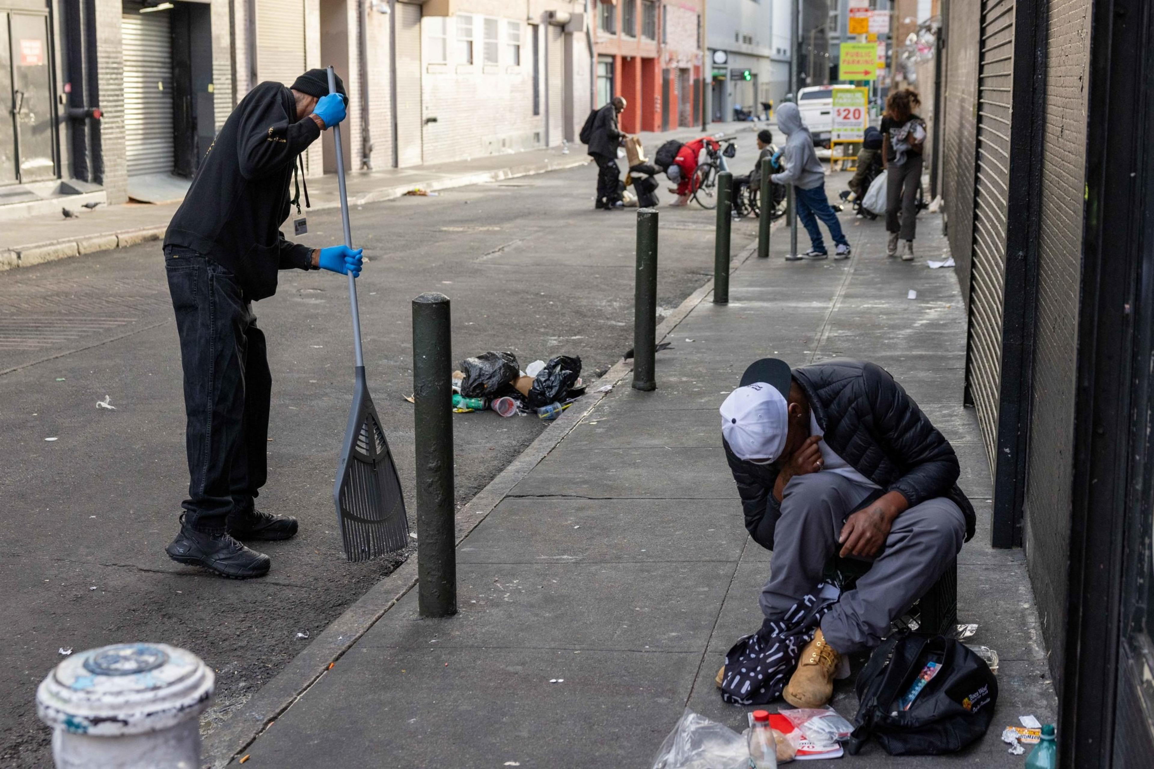 A street scene shows a person sweeping the pavement, while another sits nearby with belongings scattered around. More people and litter are visible in the background.