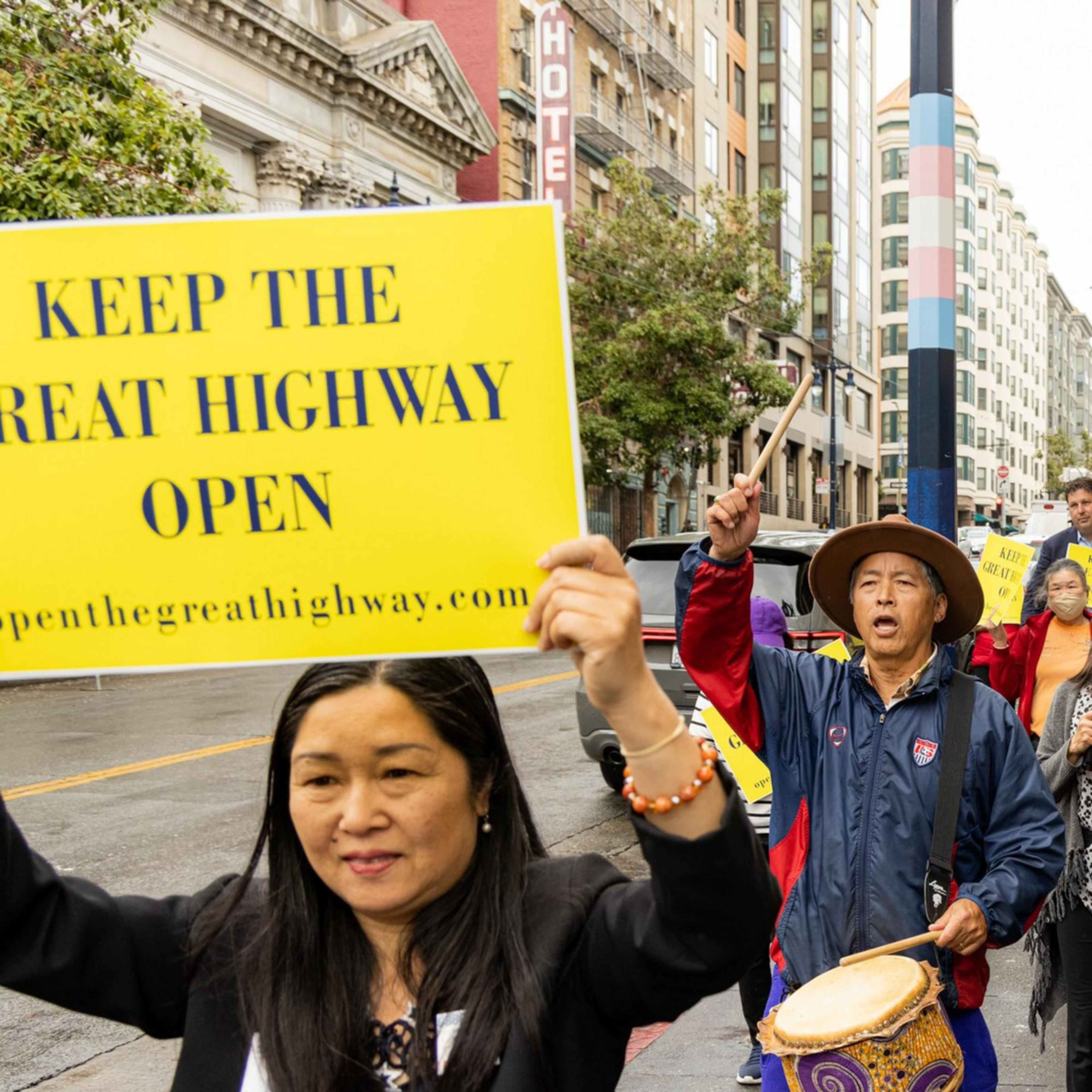 A group of people marches on a city street holding up yellow signs that read, &quot;KEEP THE GREAT HIGHWAY OPEN.&quot; One person is playing a drum.