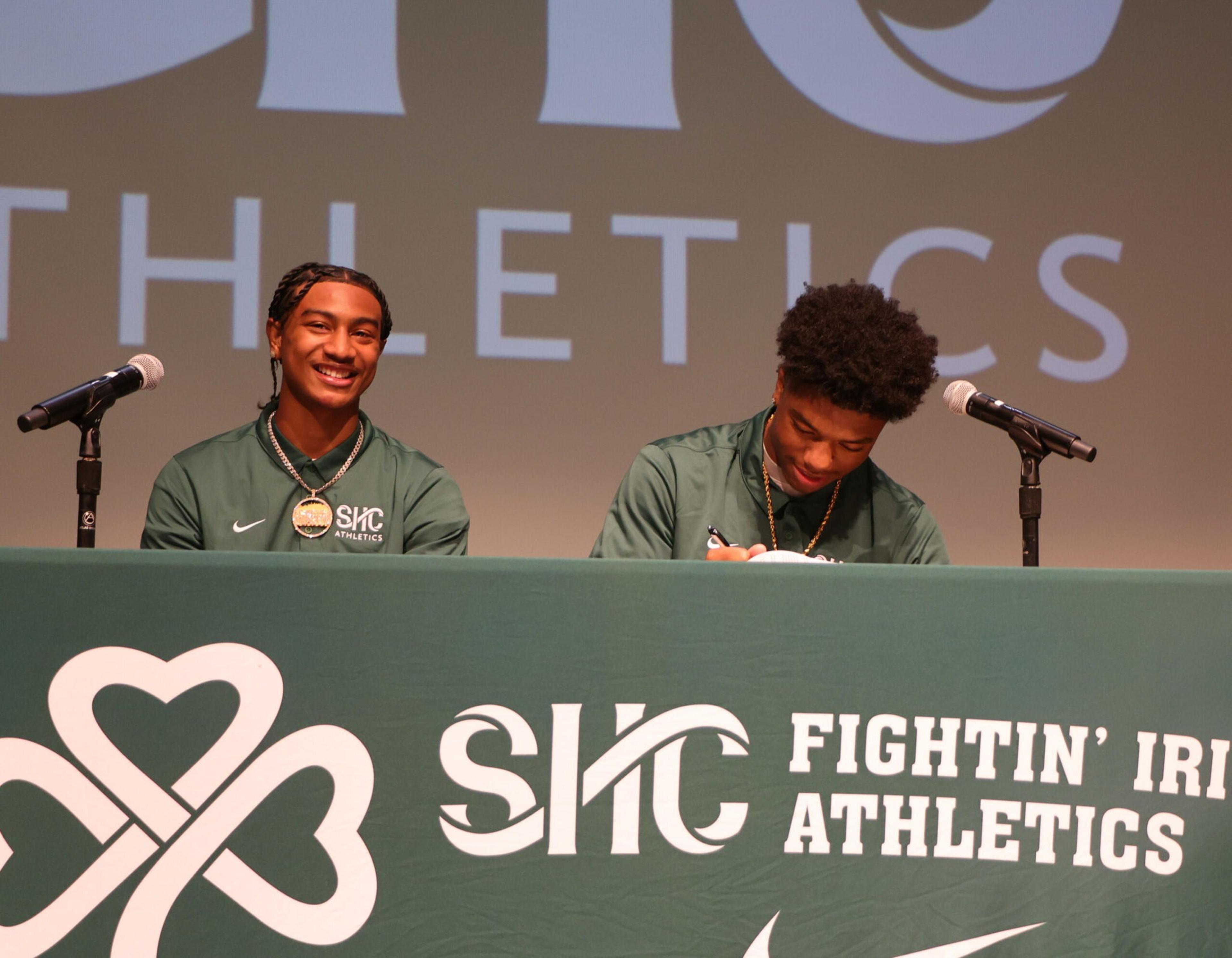 Two young men in green SIC Athletics jackets sit at a table with microphones; one smiles at the camera while the other signs a document.
