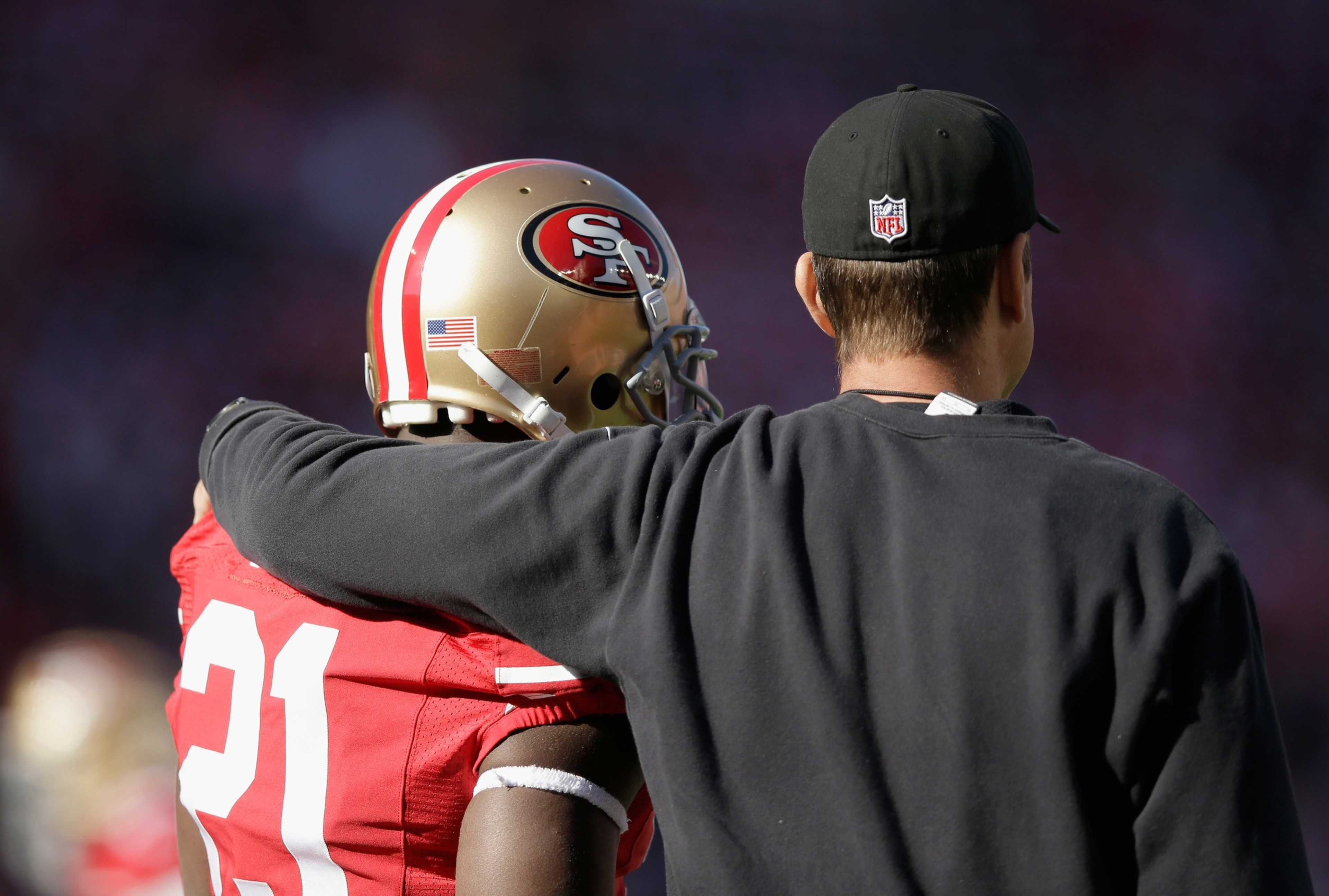 A football coach in black with an NFL cap has his arm around a San Francisco 49ers player wearing a helmet and jersey number 21.