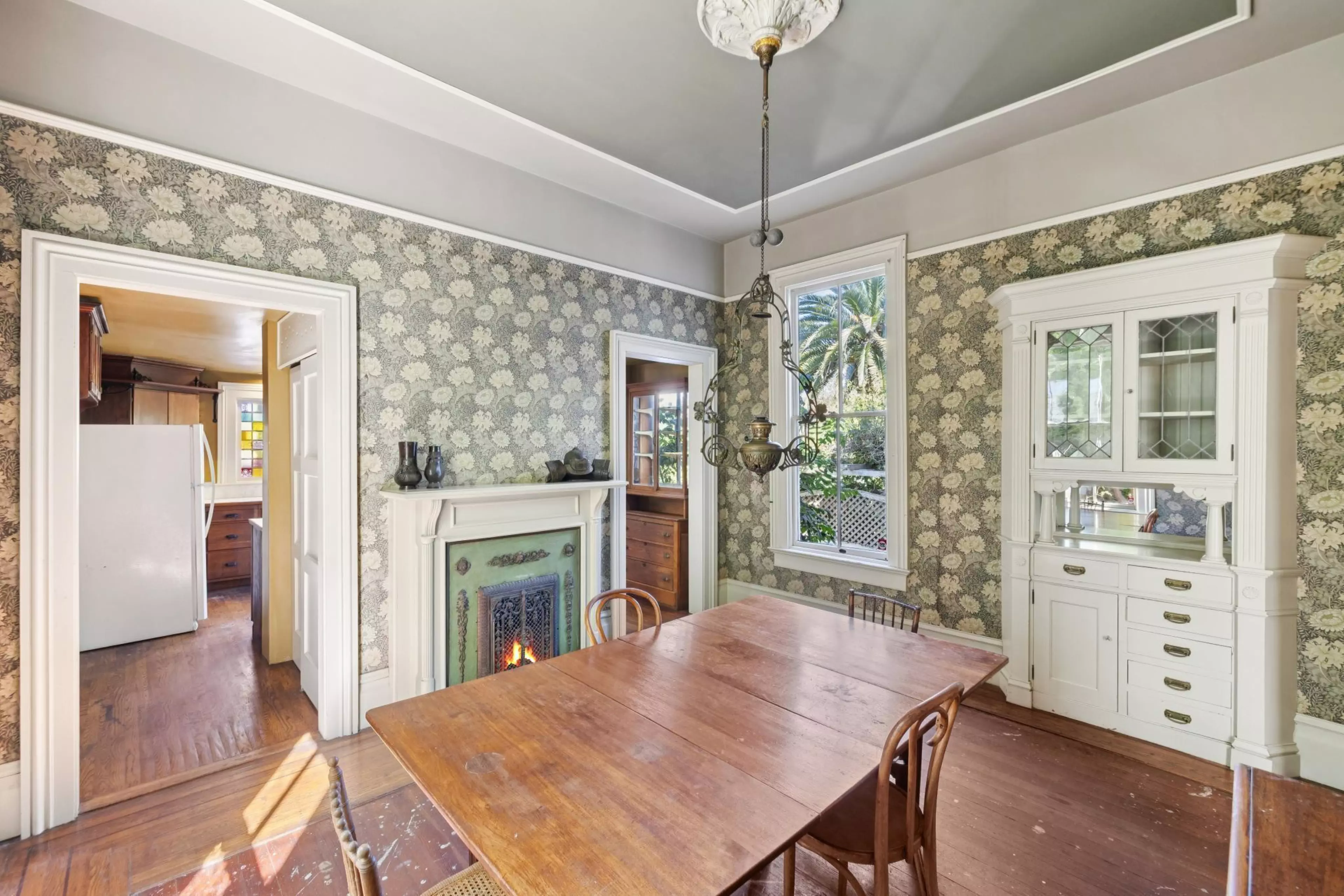 A dining room features floral wallpaper, a wooden table with four chairs, a white fireplace with a small fire, a vintage chandelier, and a white built-in cabinet.