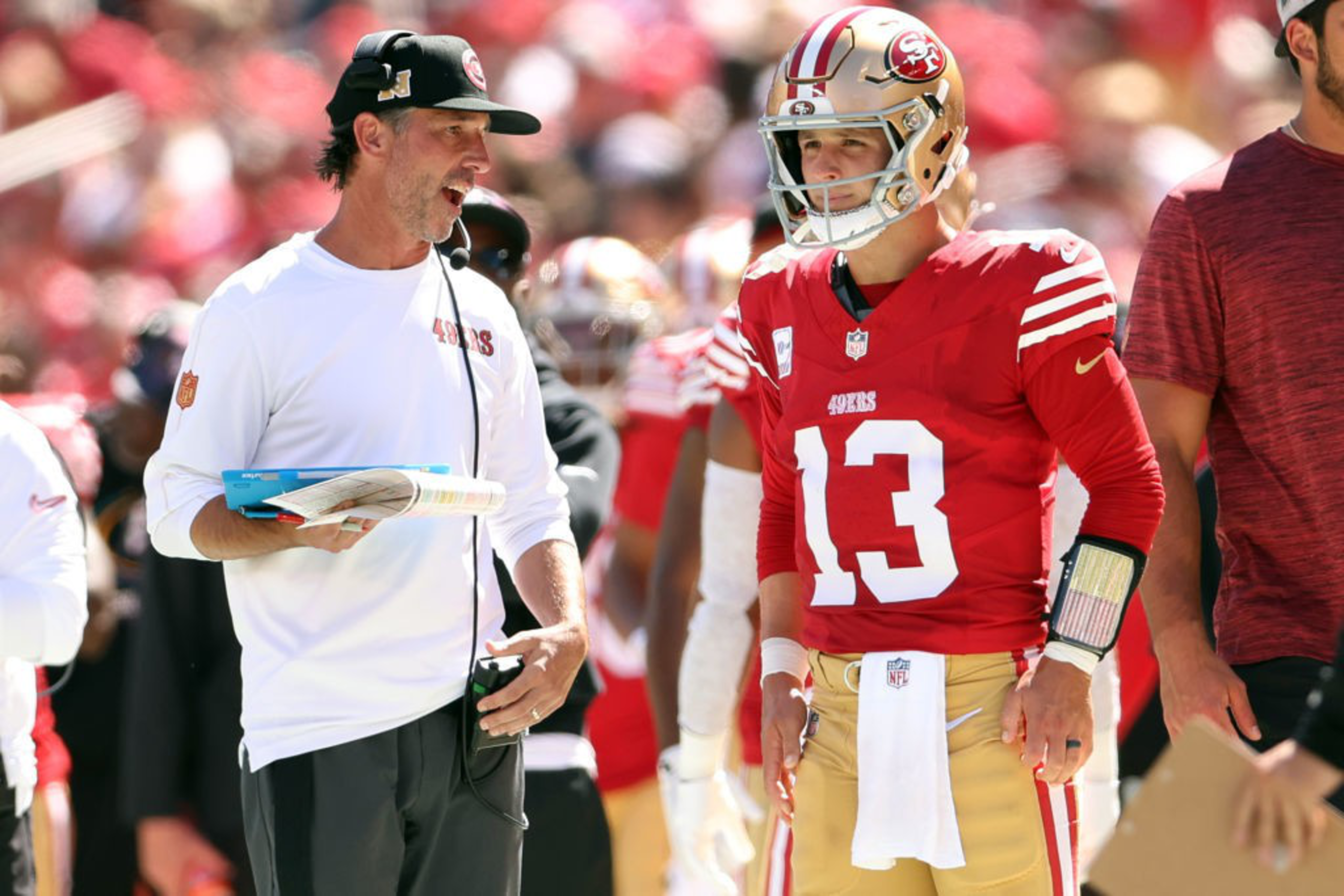 A football coach in a white shirt talks animatedly to a player in a red 49ers uniform and helmet. The player listens intently, with others in the background.