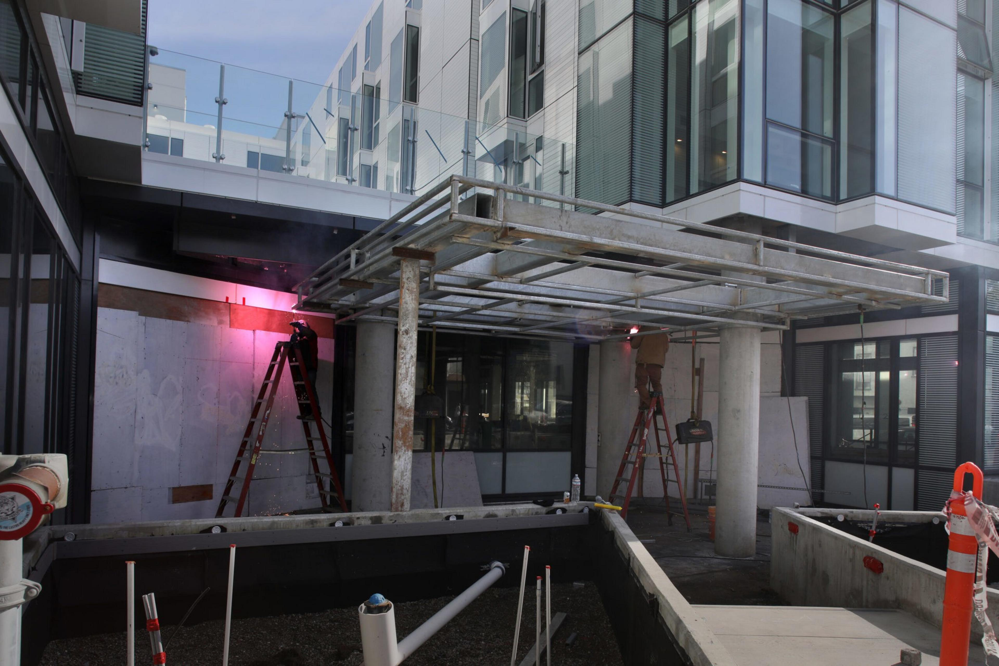 Two workers on ladders weld a metal frame structure under a building overhang, with pipes and construction materials in the foreground.